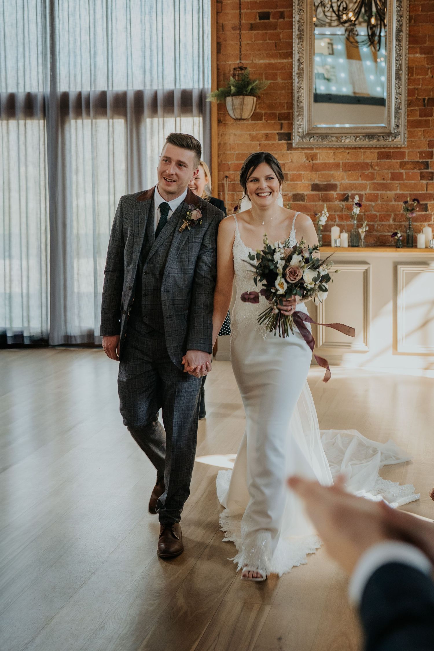Smiling newlyweds walk hand-in-hand through a stylish brick-walled venue, bride carrying a beautiful bouquet.