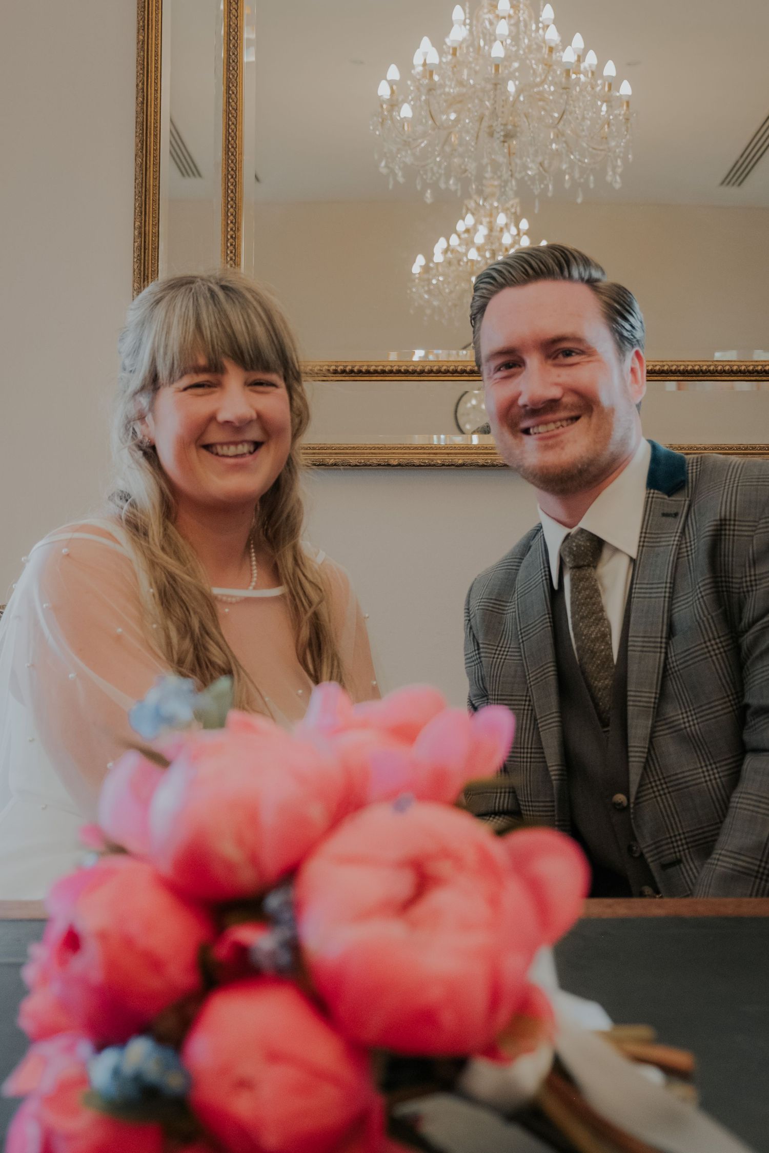 Smiling bride and groom sit at signing table with pink peony bouquet, elegant chandelier backdrop.