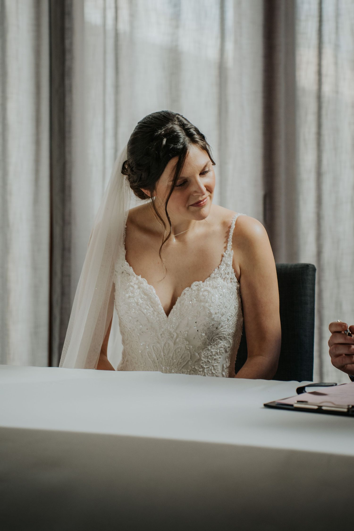 Smiling bride in lace wedding dress and veil sits at a white table near sheer curtains on her wedding day.