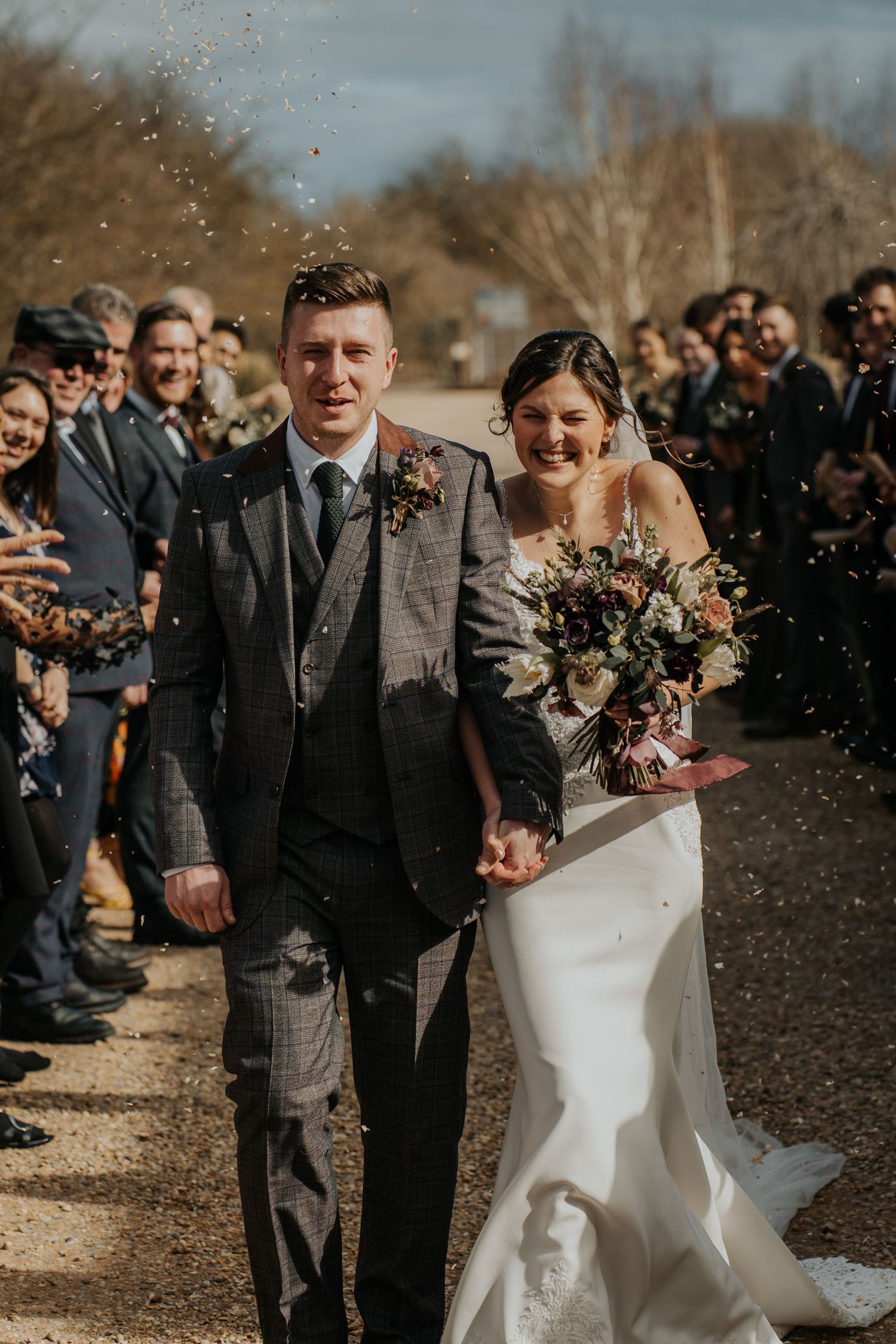Newlywed couple walking through confetti shower, bride holding bouquet, groom in plaid suit, guests cheering outdoors.