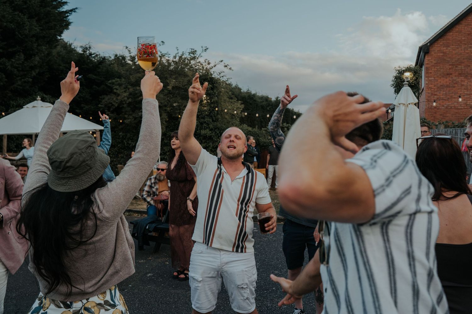 Group of friends dancing outdoors at a summer party, raising glasses in celebration near a pub garden.