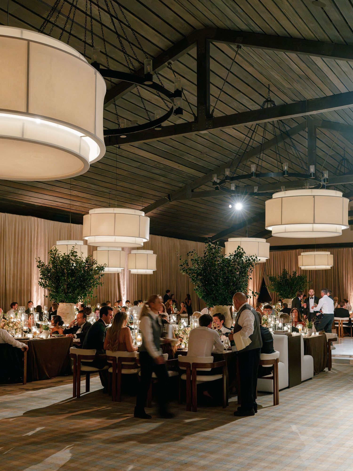 Elegant wedding reception hall with large drum pendant lights, greenery centerpieces, and guests dining at long tables.