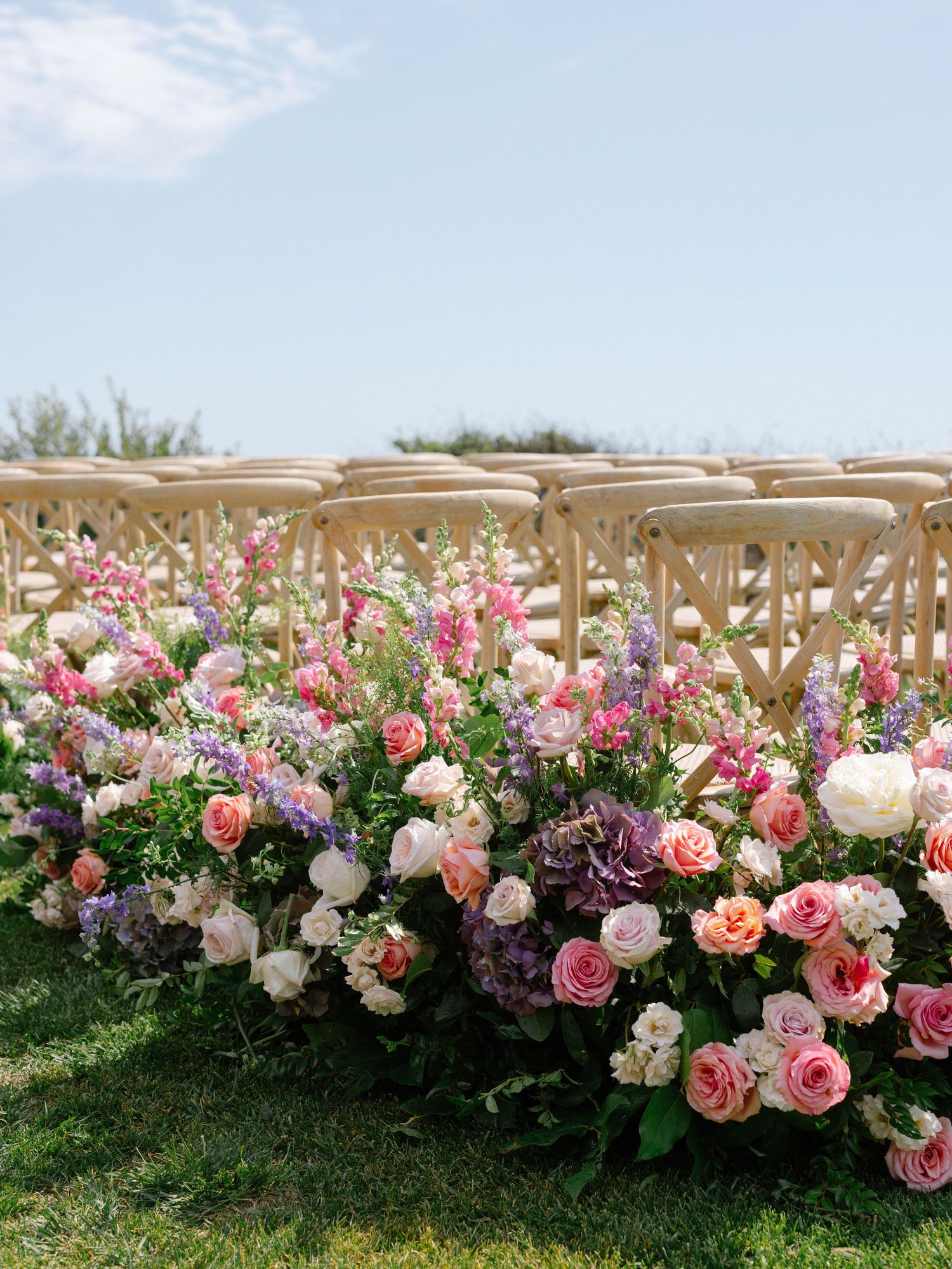 Lush garden-style wedding ceremony aisle flowers featuring pink, purple, and white roses with wooden chairs outdoors.