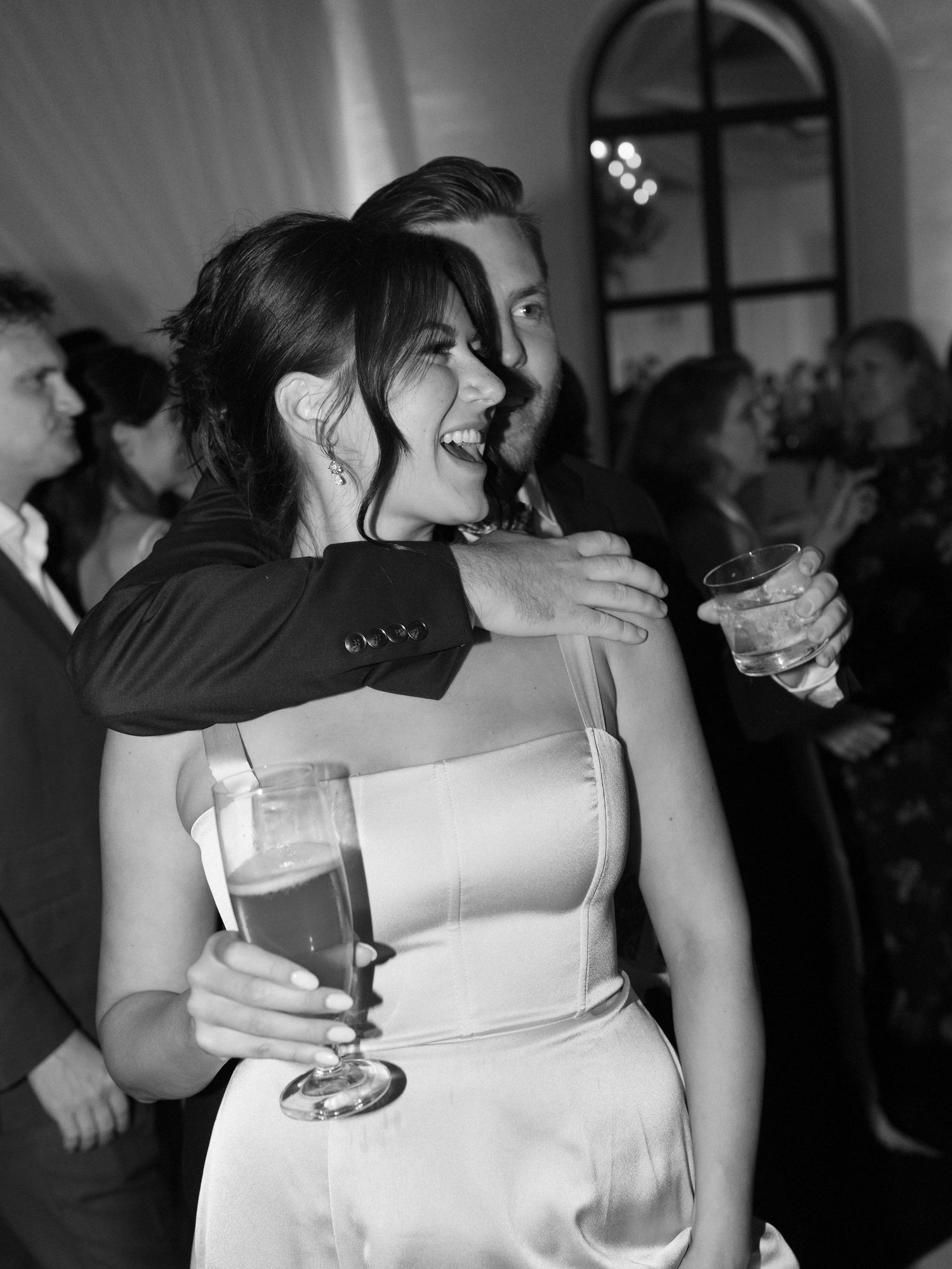 Joyful wedding guests laugh and celebrate, woman in white dress holding champagne flute at reception.