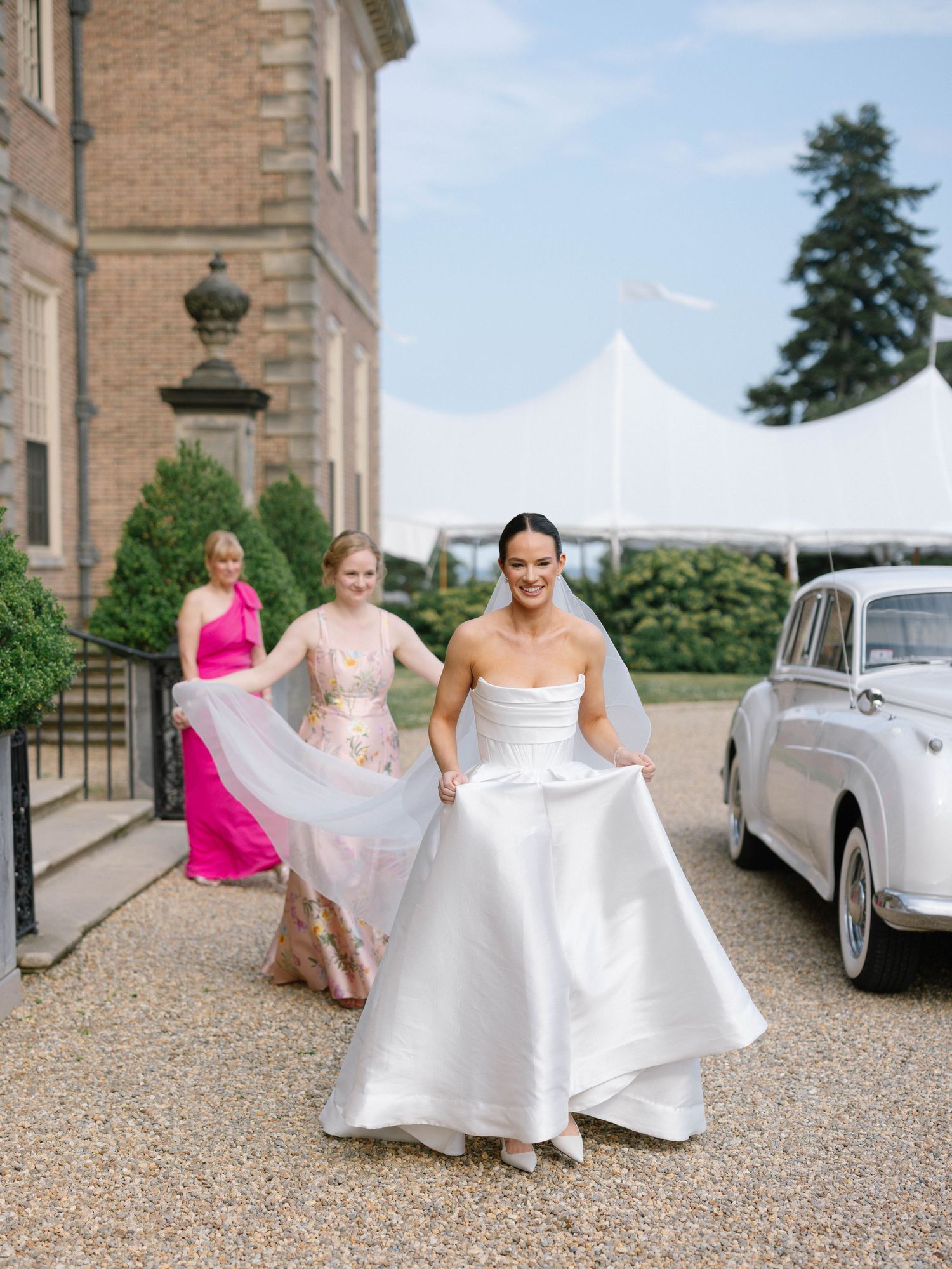 Bride in a strapless white ballgown walks briskly toward a classic white car accompanied by bridesmaids in pink.