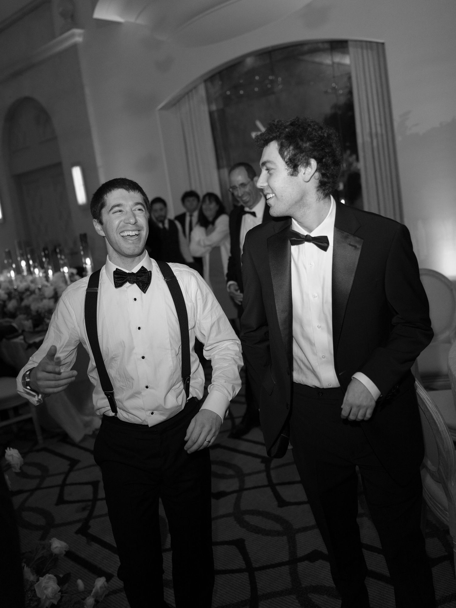 Two men in tuxedos laughing together on elegant wedding reception dance floor.