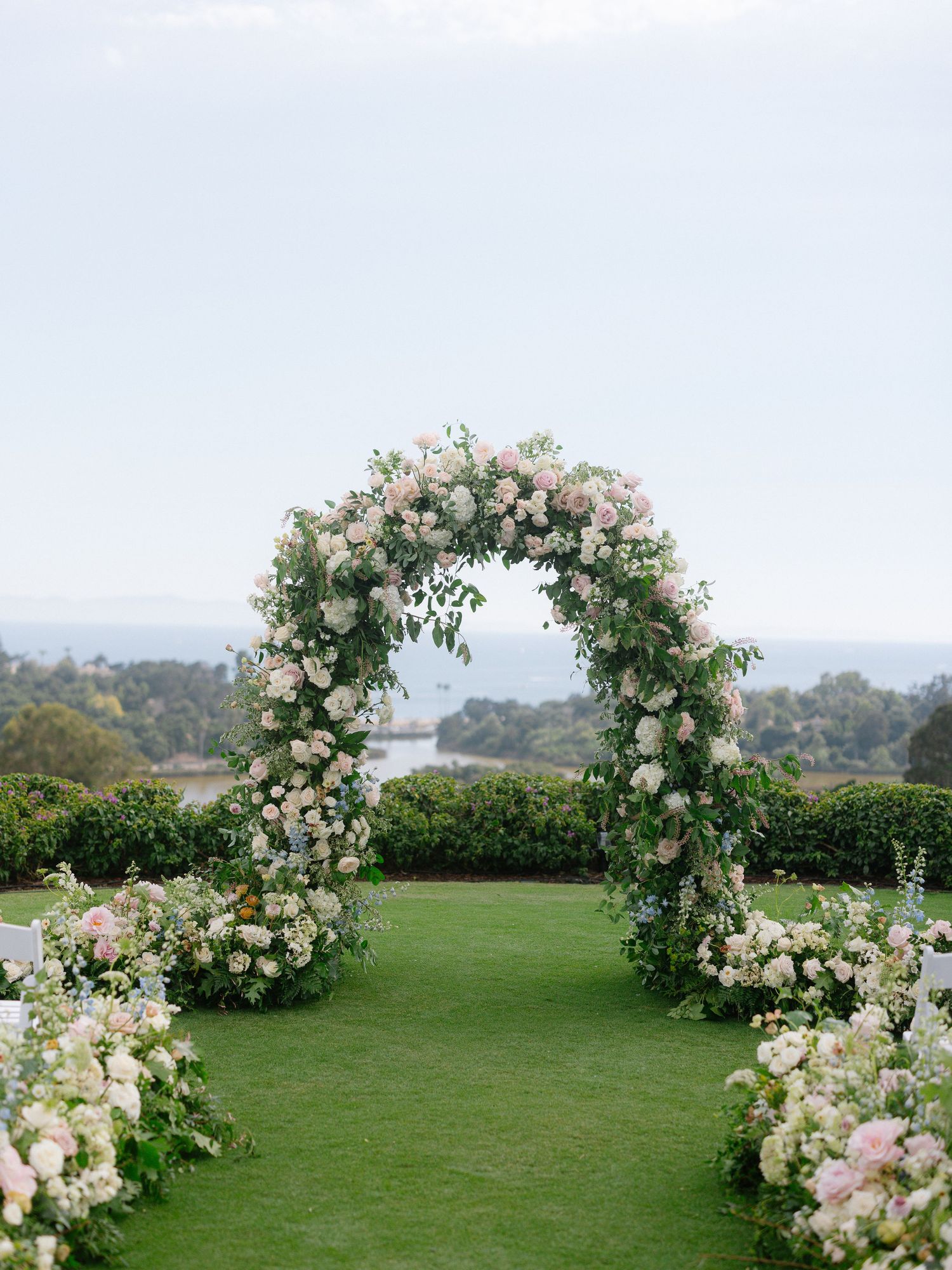 Romantic floral arch with pink and white blooms frames ocean view at outdoor wedding ceremony.