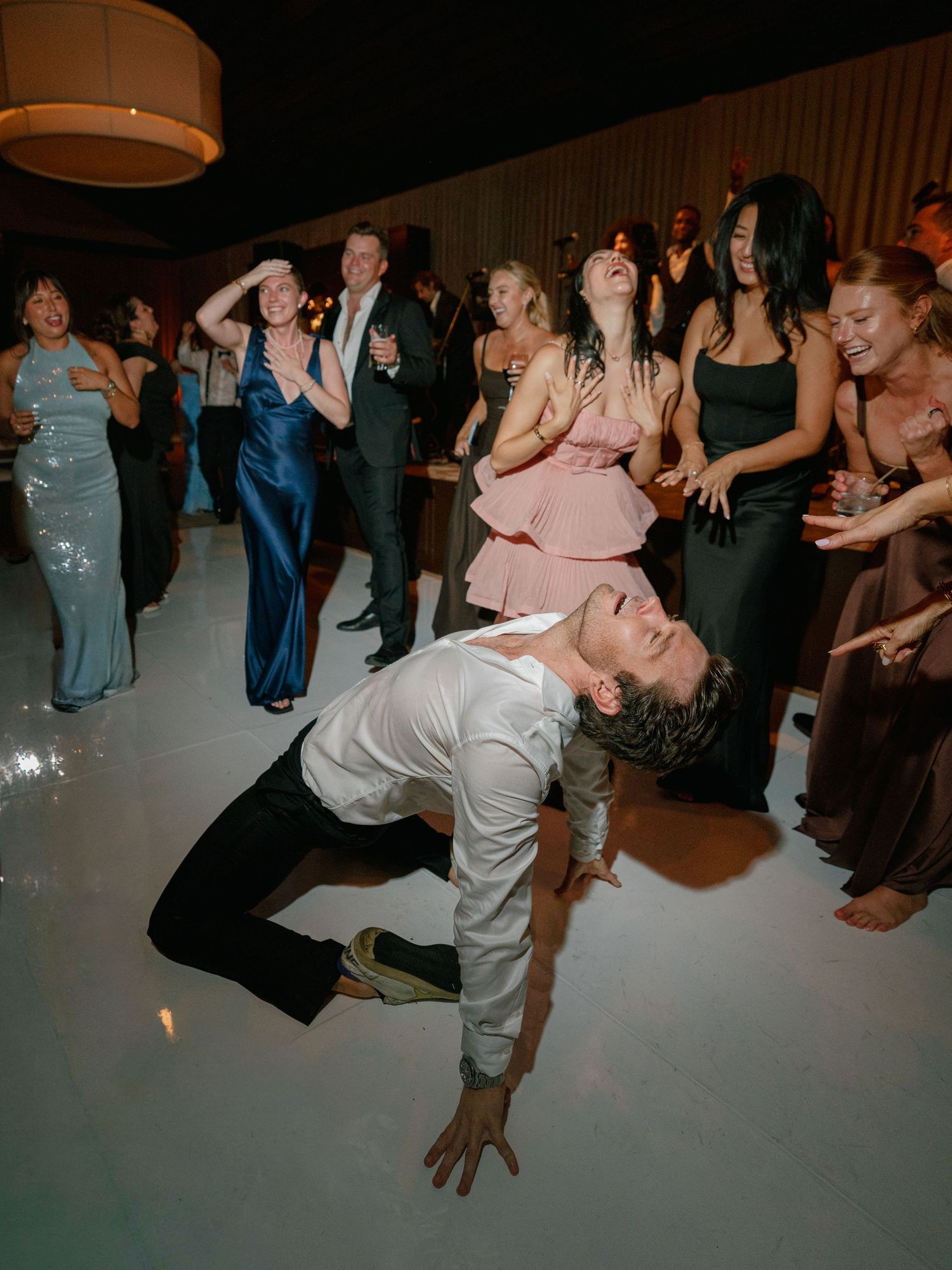 Wedding guest performing dramatic dance move on white dance floor surrounded by laughing guests.