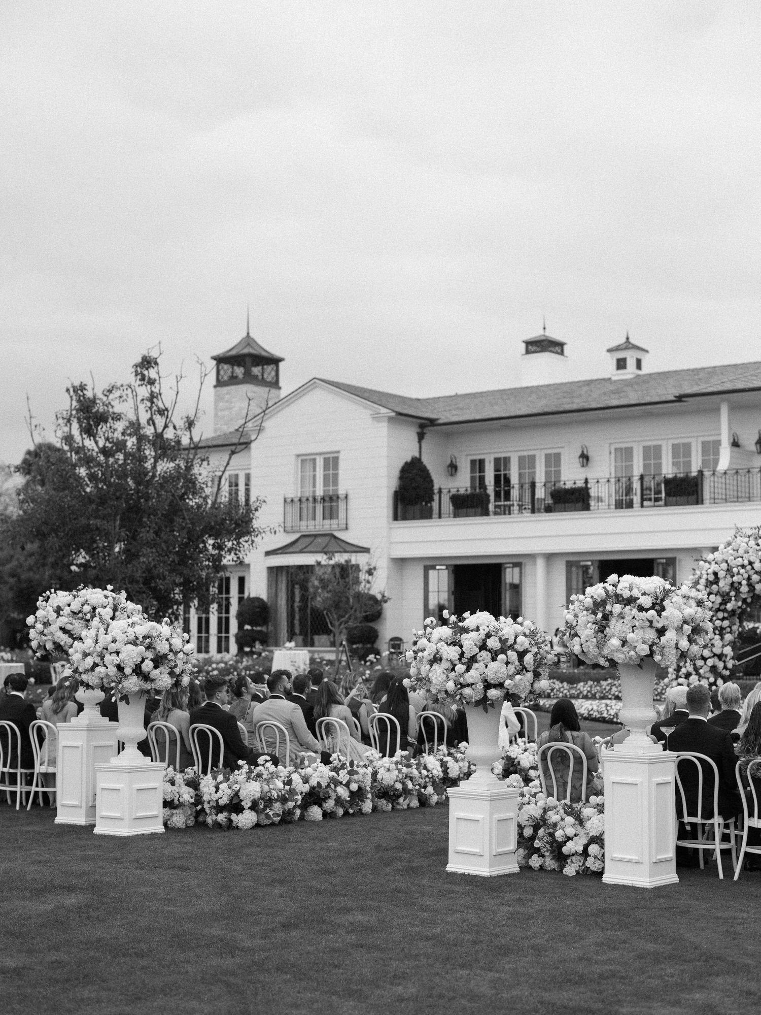 Black and white photo of outdoor wedding ceremony with lush floral arrangements before grand white estate.
