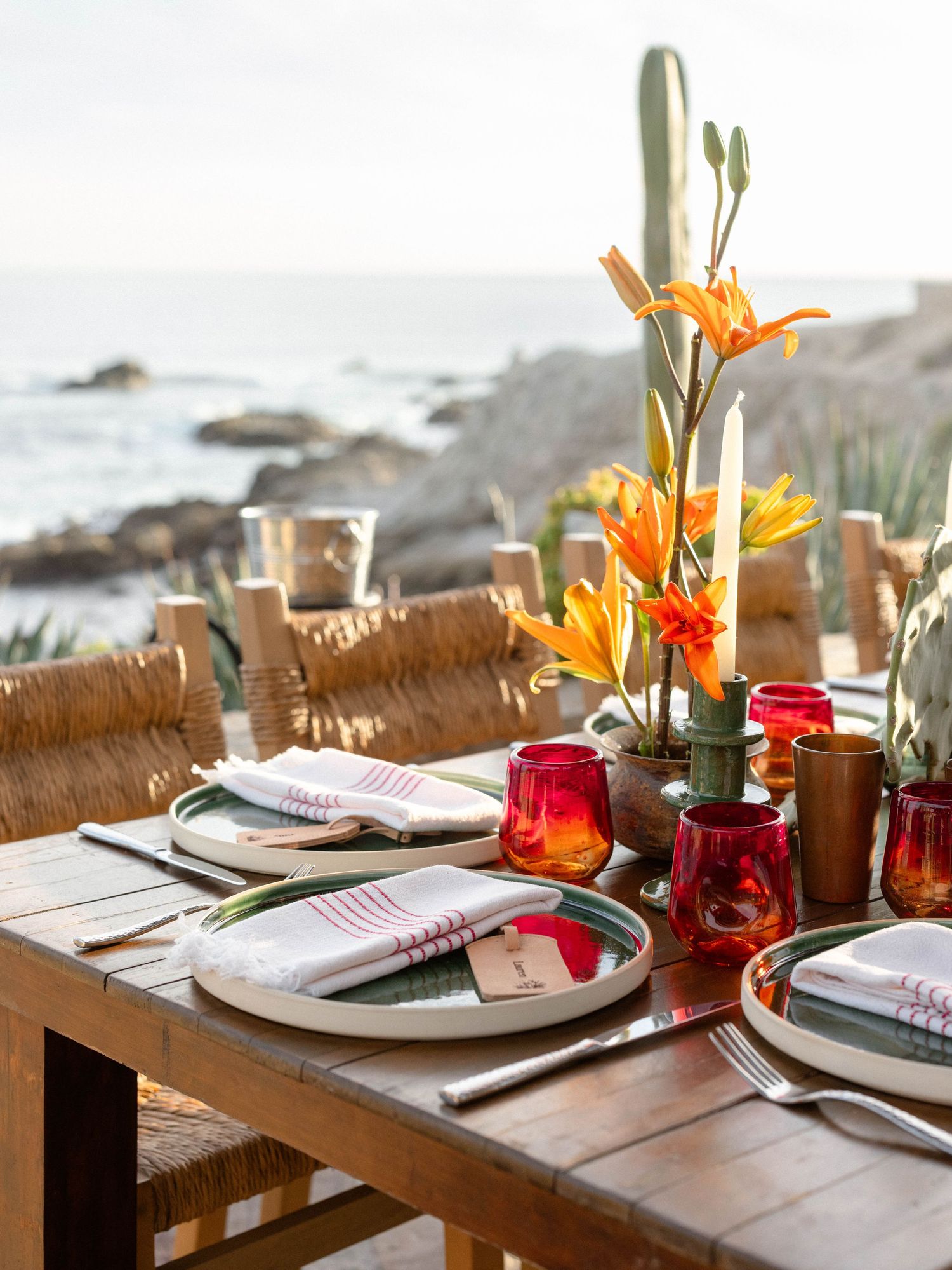 Rustic wooden outdoor dining table with orange lily centerpiece, red glassware, and ocean view at sunset.