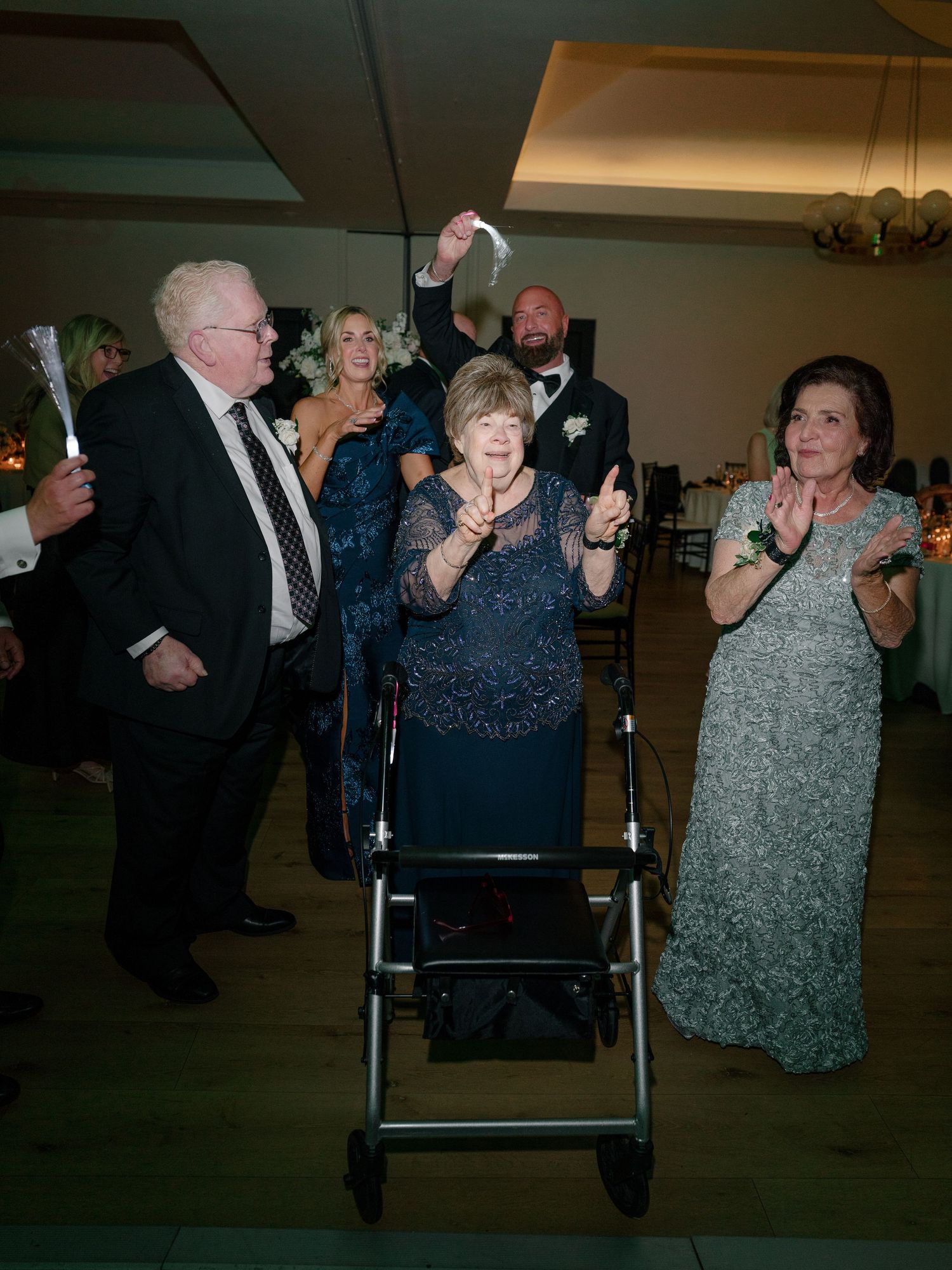 Elderly woman with walker celebrating at wedding reception surrounded by family clapping and cheering on dance floor.