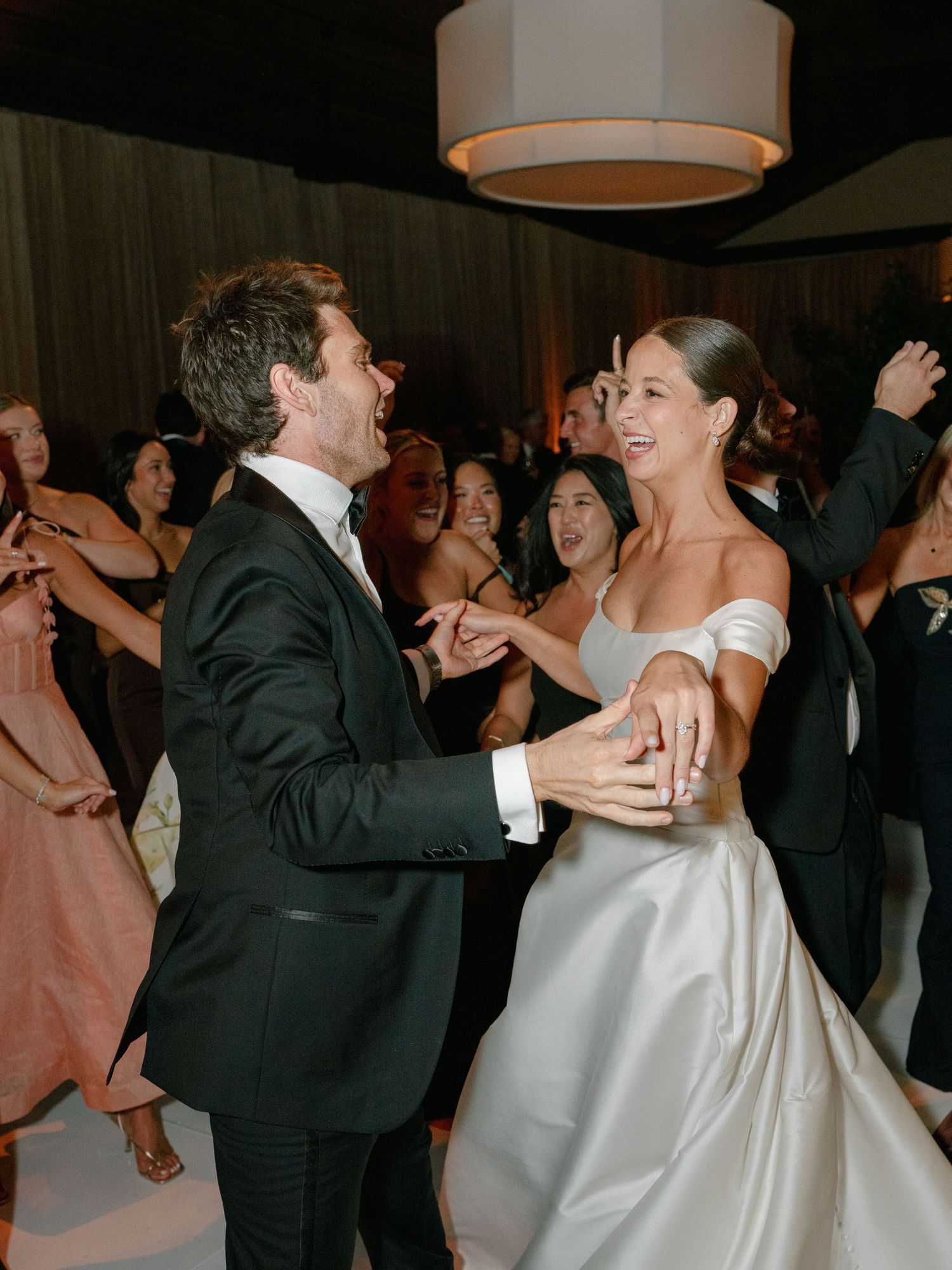 Bride in off-shoulder white gown and groom in black tuxedo dancing joyfully at wedding reception.