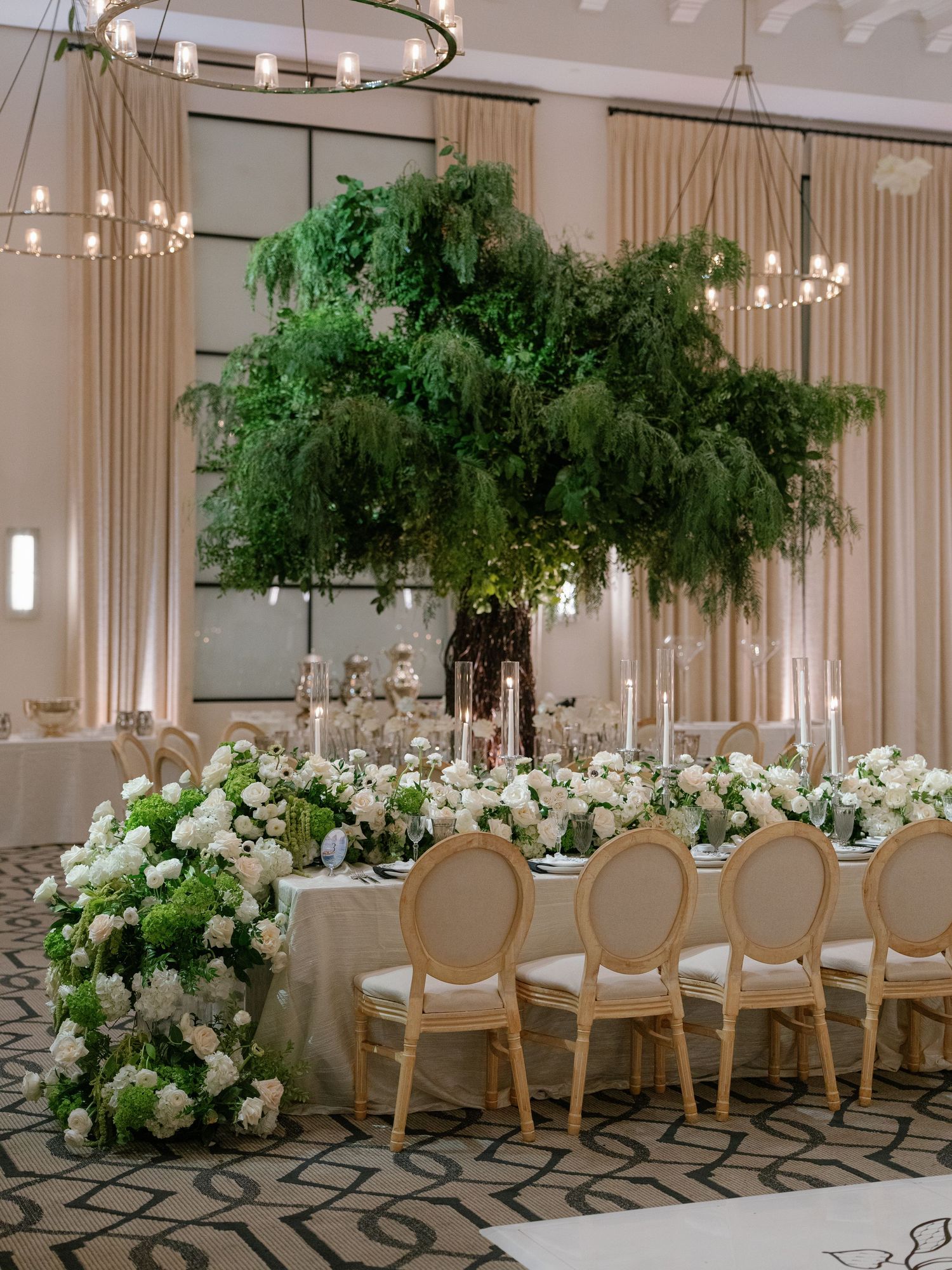 Elegant wedding reception with lush green hanging floral installation above a long white table with white flower arrangements.