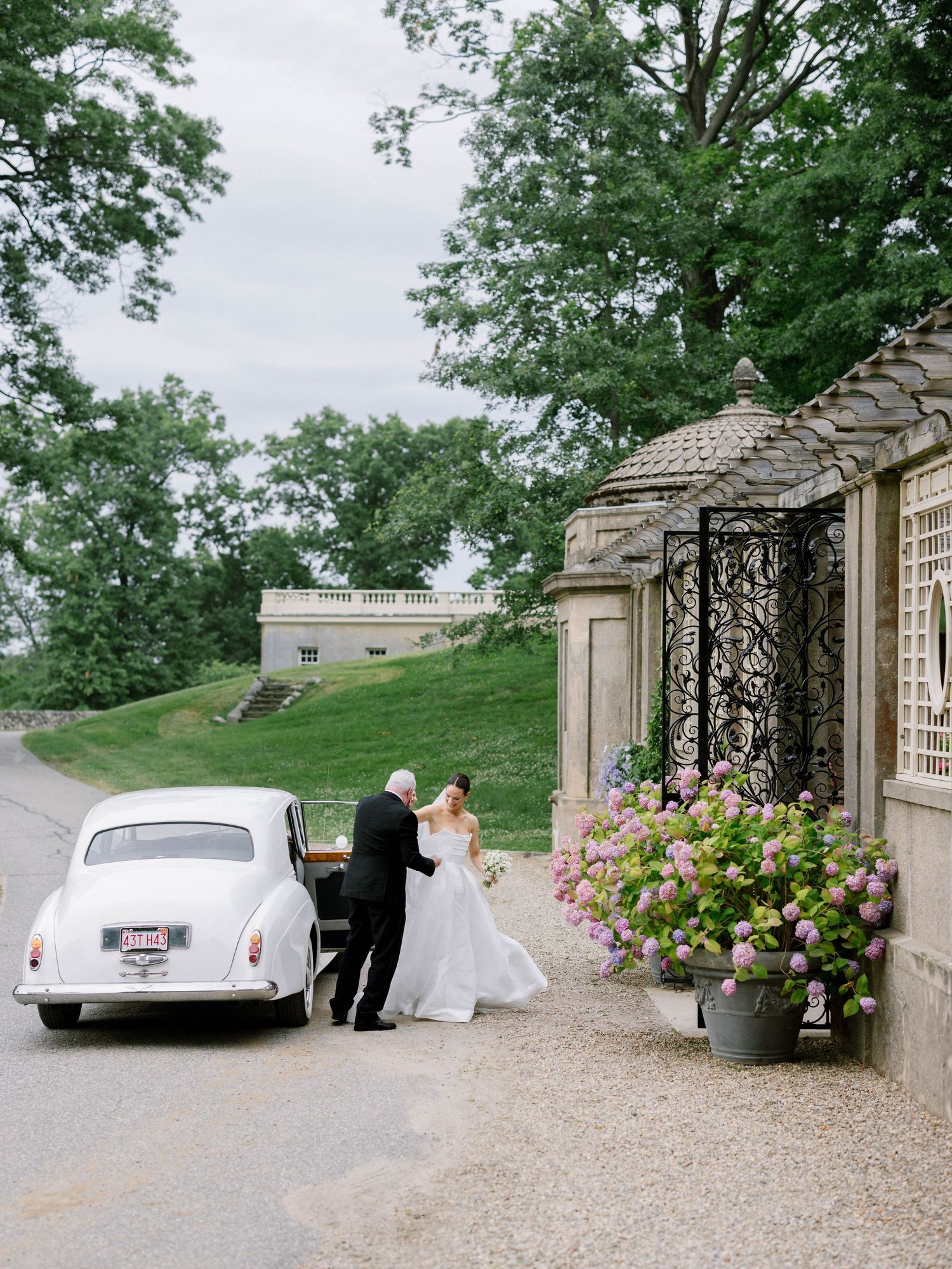 Bride arriving in a vintage white car at an elegant estate wedding venue with ornate iron gates and hydrangeas.