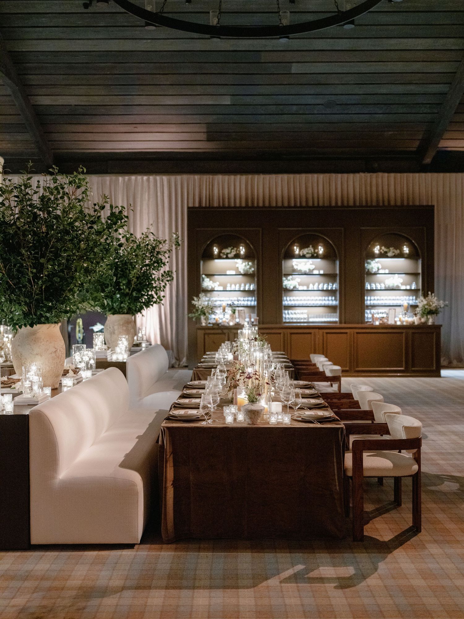 Sophisticated wedding reception with white banquette seating, long dining tables, and illuminated bar cabinet backdrop.