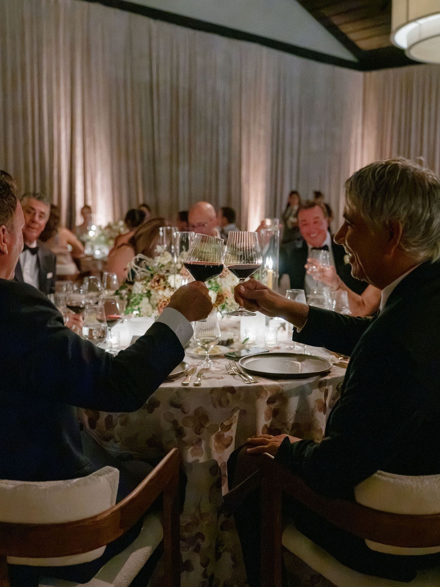 Wedding guests clinking red wine glasses in toast at candlelit reception dinner table.