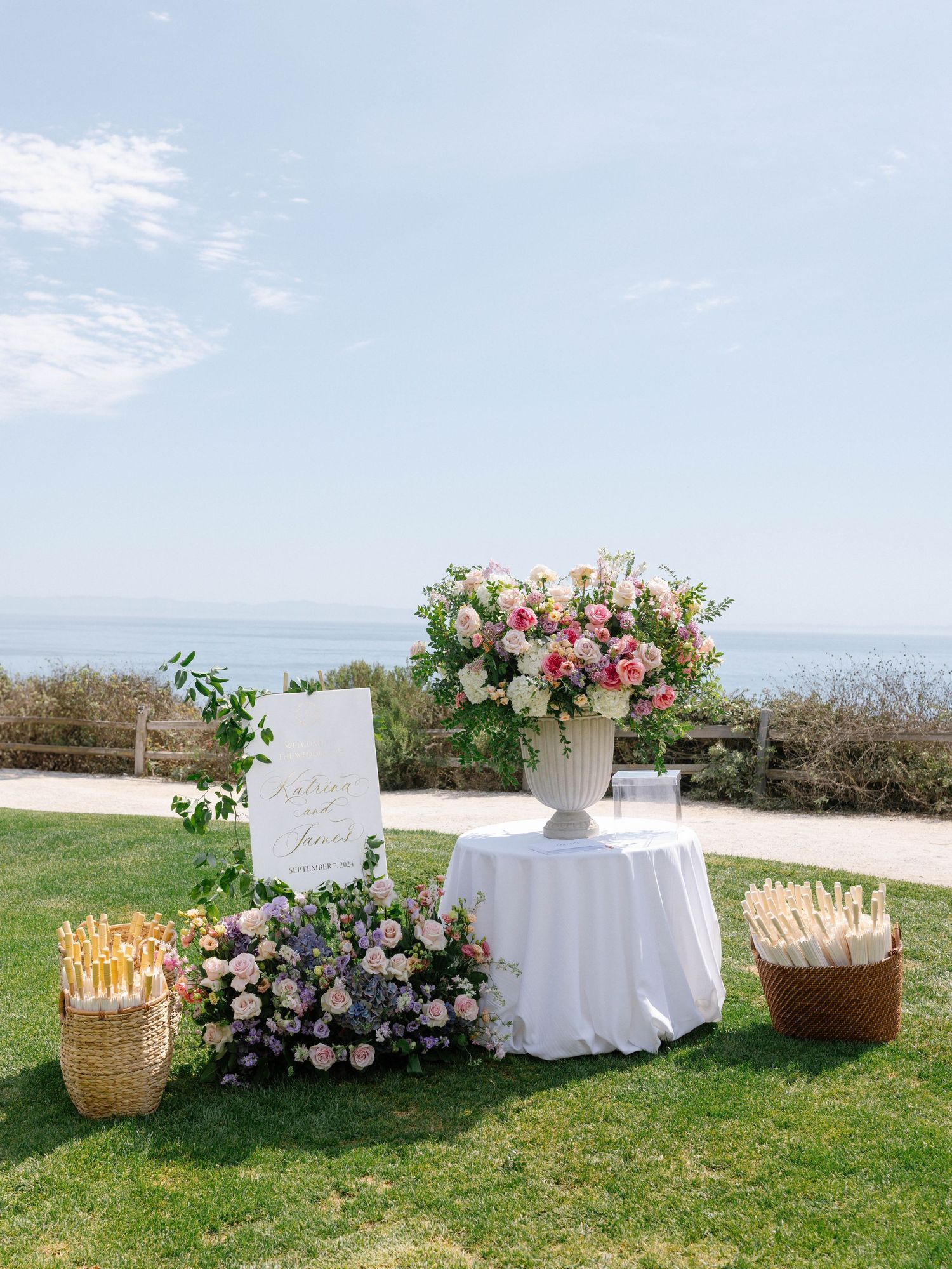 Elegant outdoor wedding welcome table with large floral arrangement, pink roses, and ocean backdrop on sunny day.