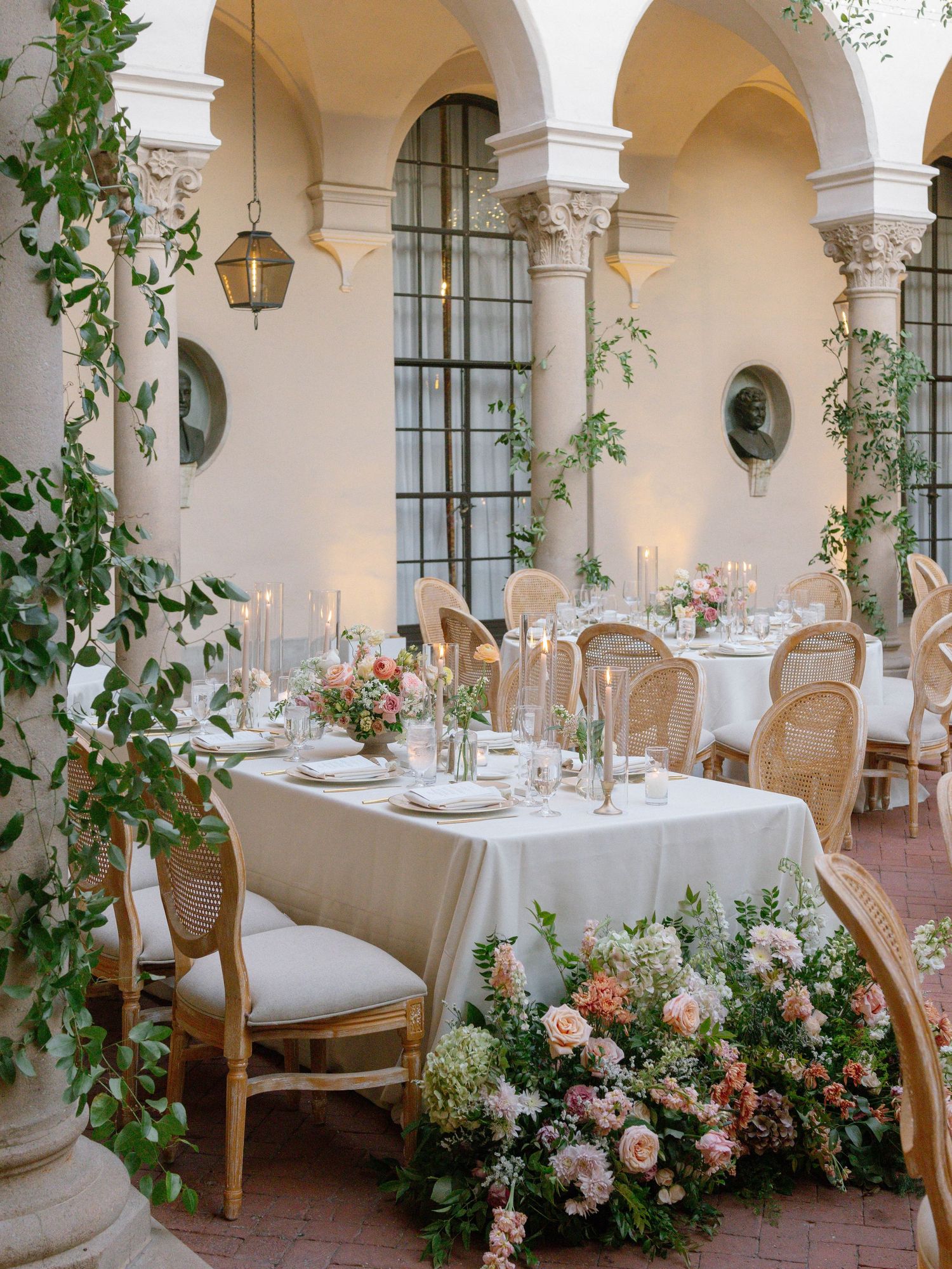 Elegant wedding reception in a courtyard with floral centerpieces, candlelight, and lush greenery on columns.