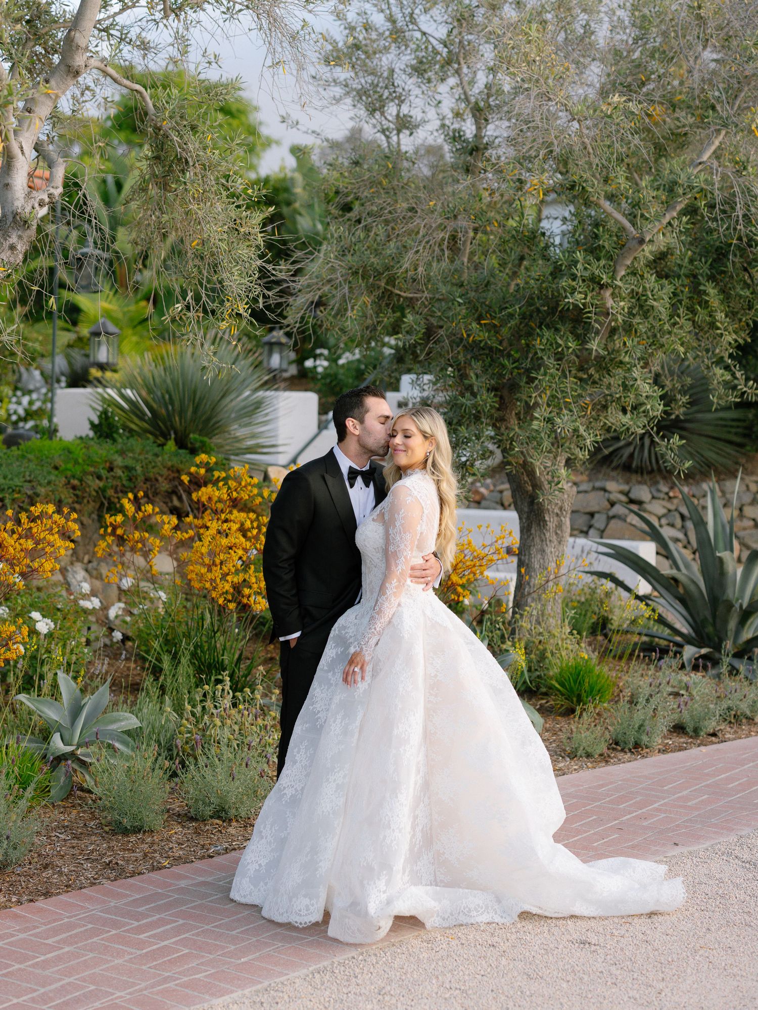 Bride in lace ballgown and groom in tuxedo share a kiss among lush garden greenery and yellow flowers.