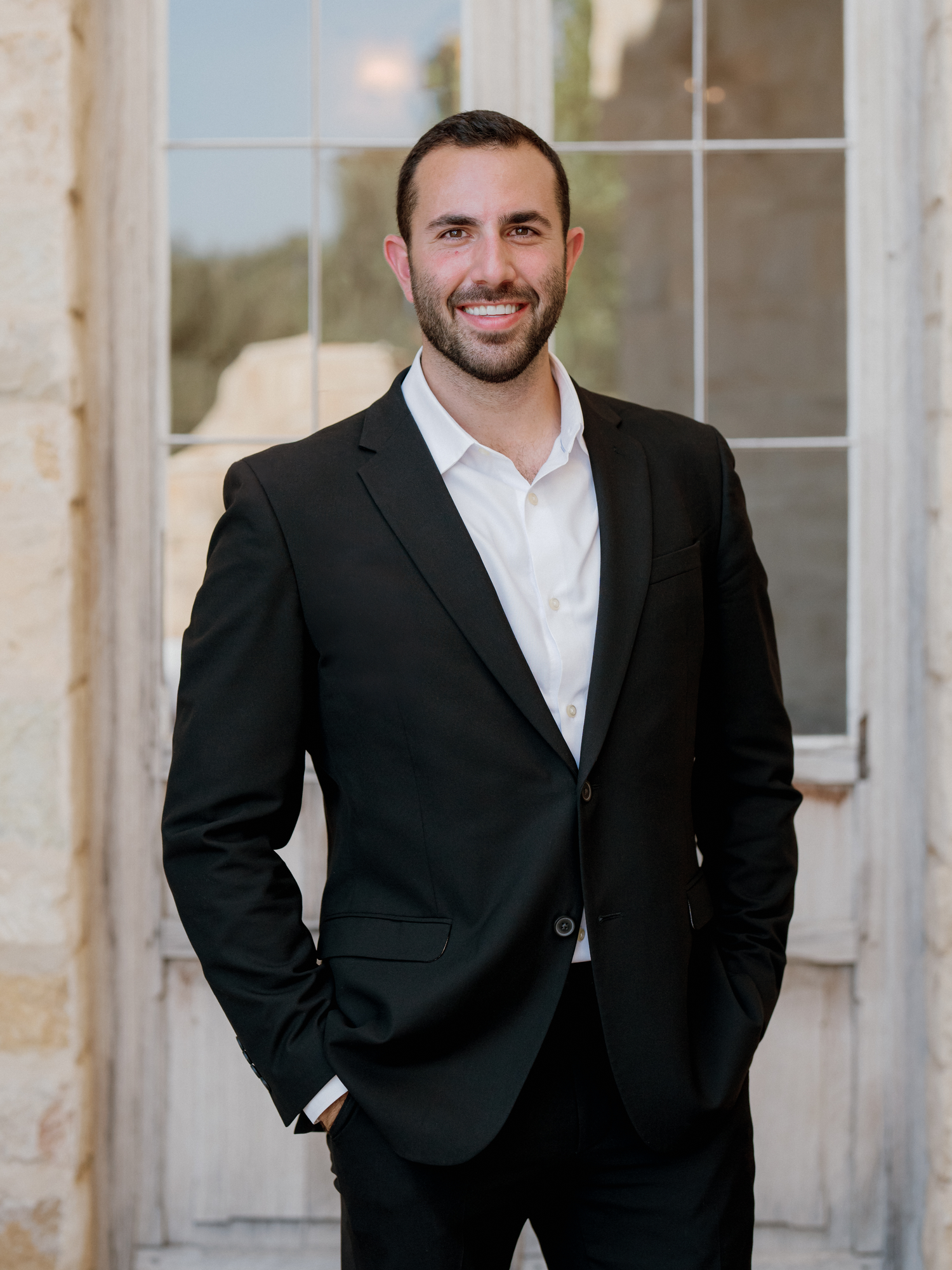 A smiling young man in a black suit and white dress shirt poses confidently outdoors near elegant stone architecture.