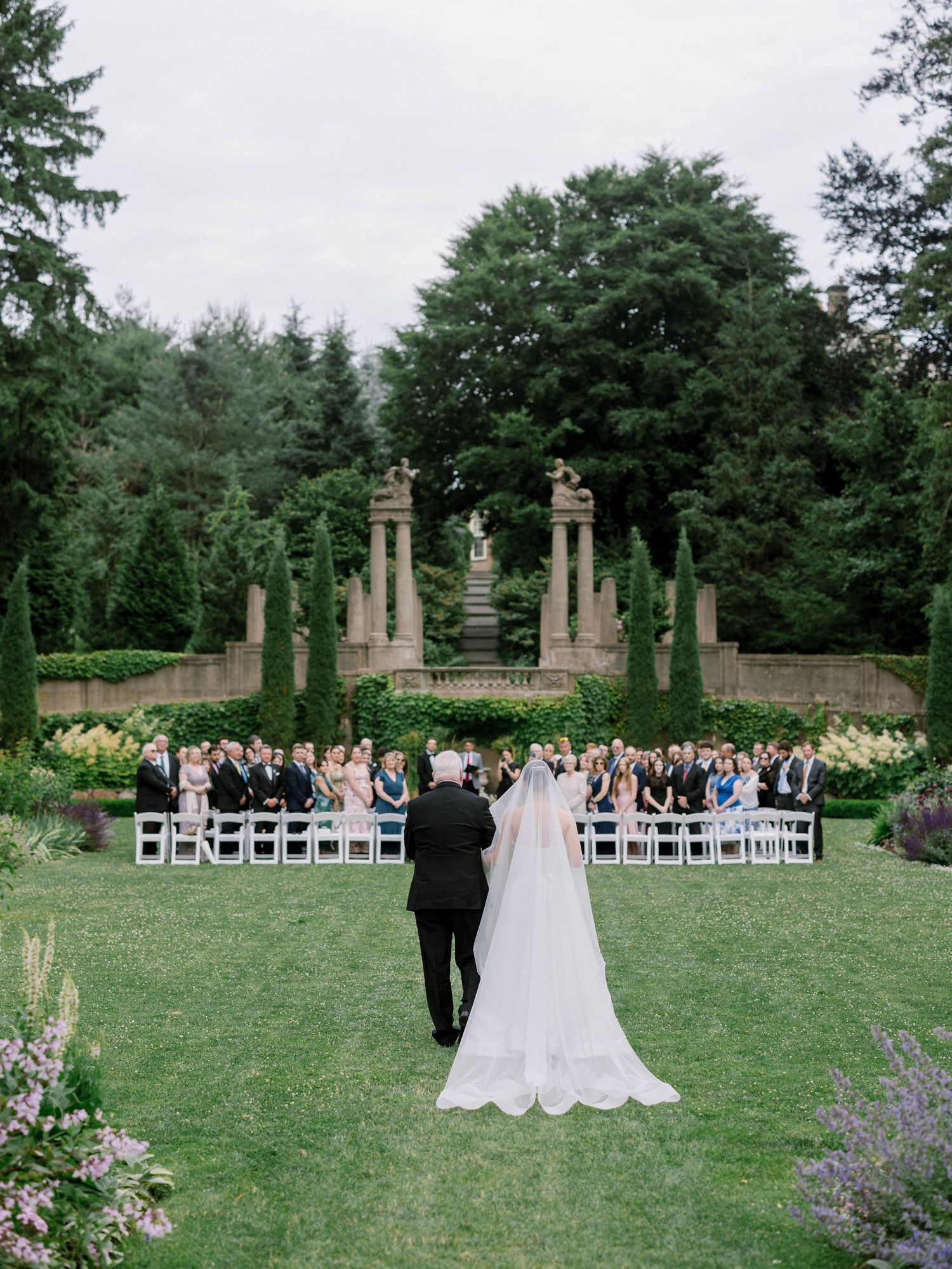 Bride in cathedral veil walks with escort toward garden ceremony with stone monument backdrop.
