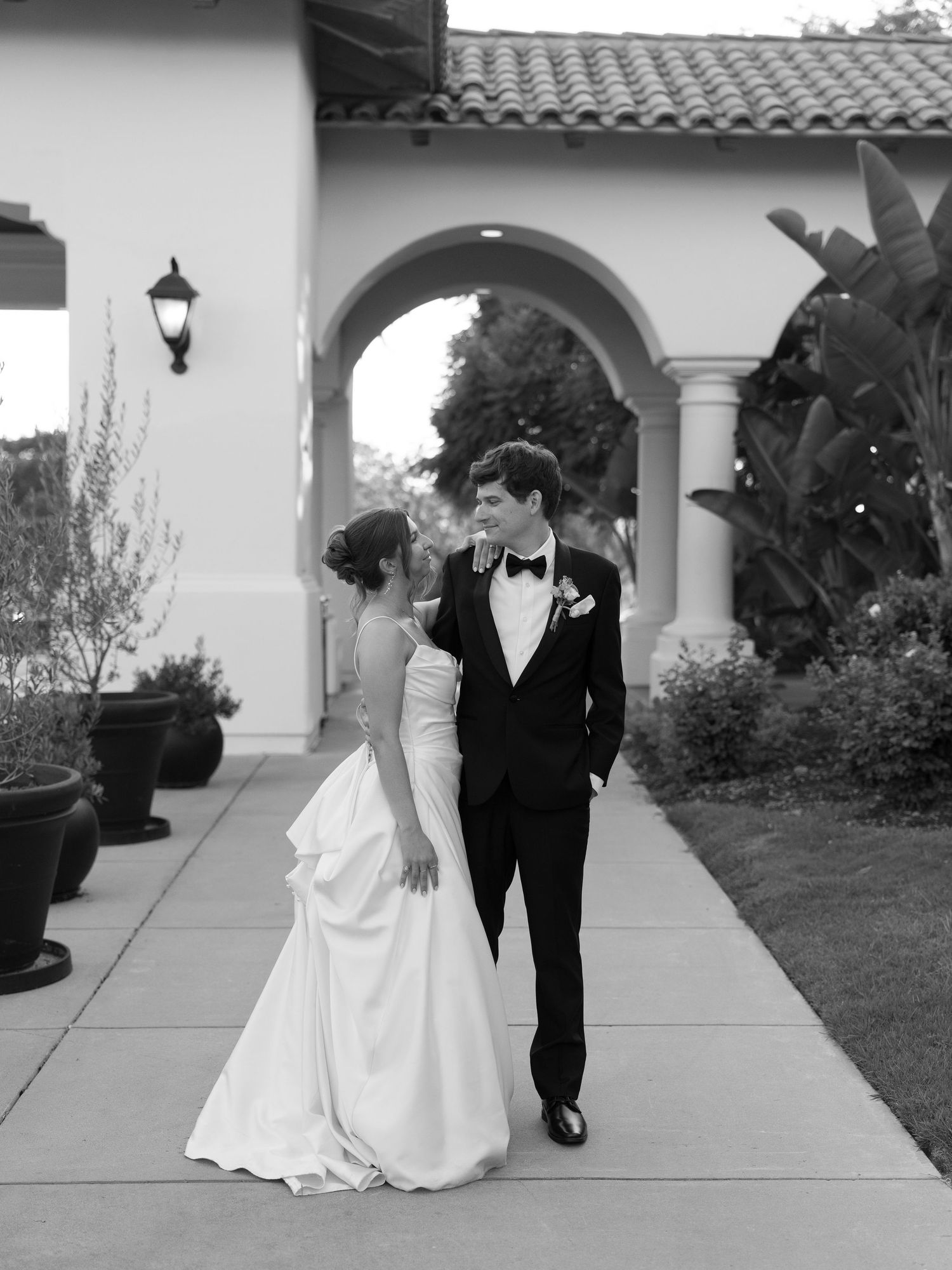Black and white photo of bride and groom walking under arched colonnade.
