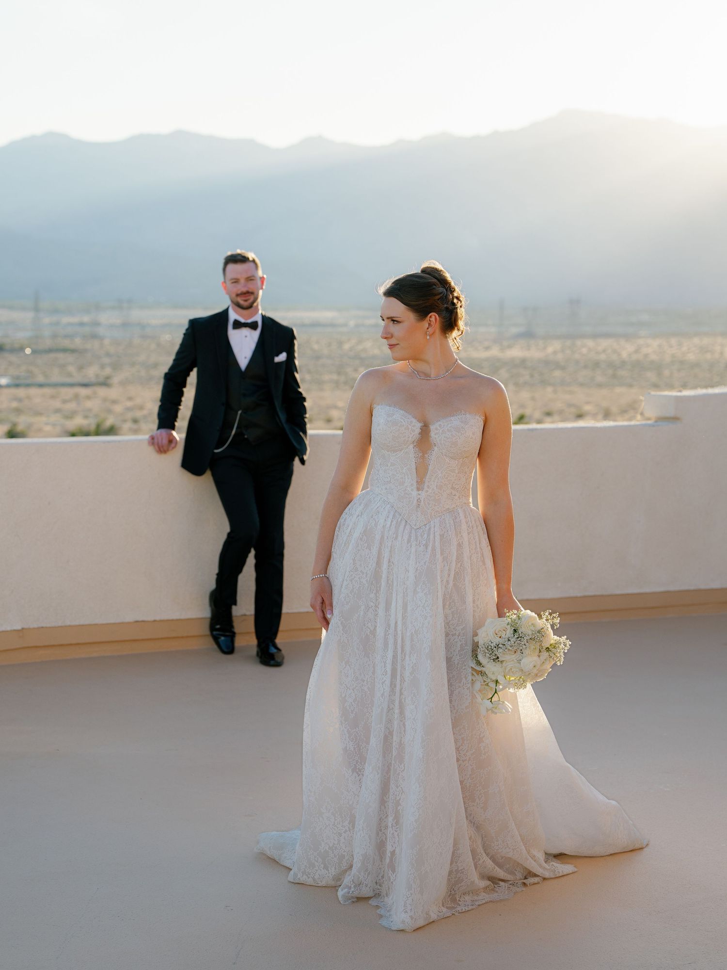 Bride in lace gown holds bouquet while groom leans against wall with stunning desert mountain backdrop.