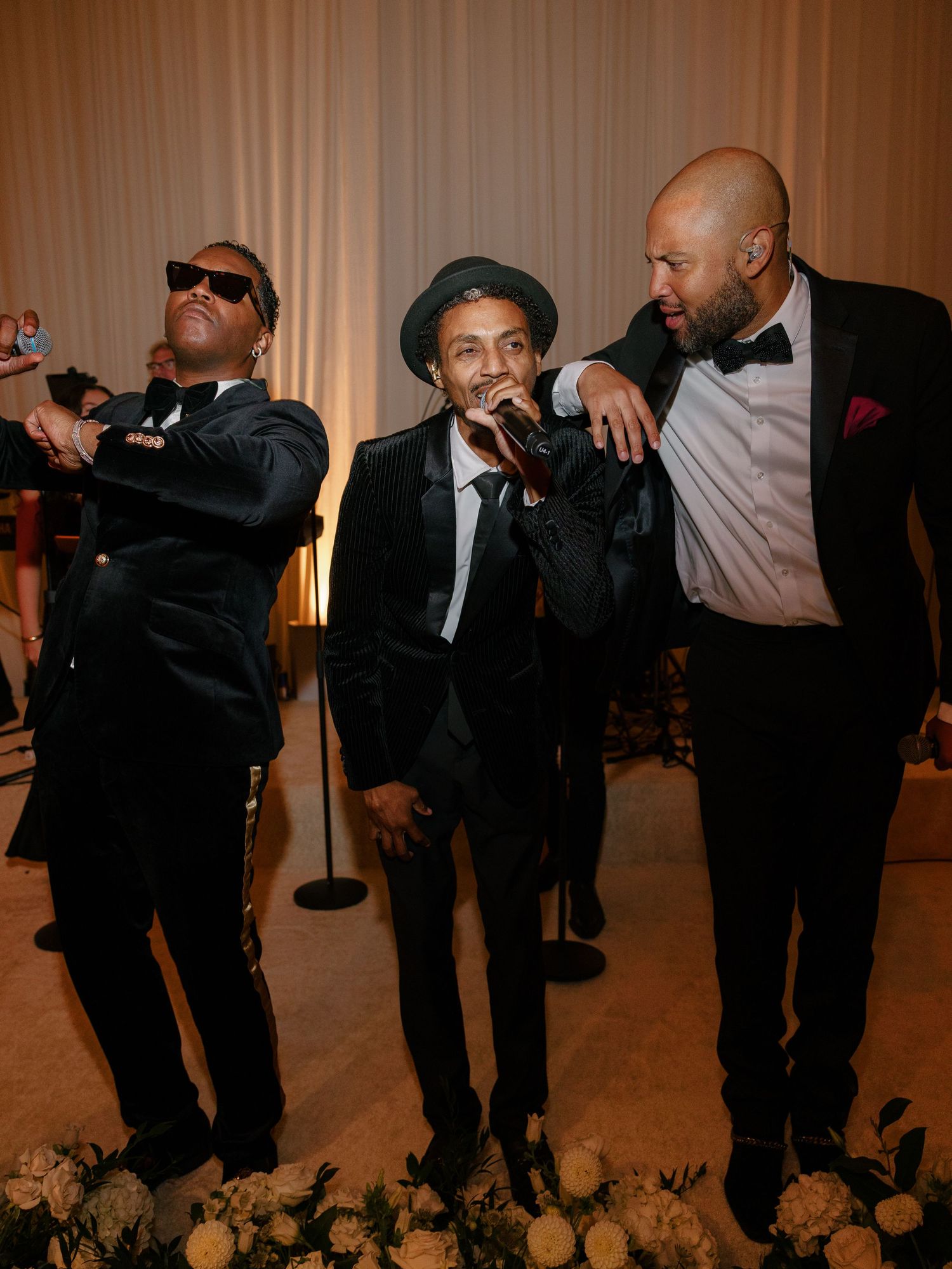 Three men in tuxedos performing and celebrating together at an elegant wedding reception with floral arrangements.
