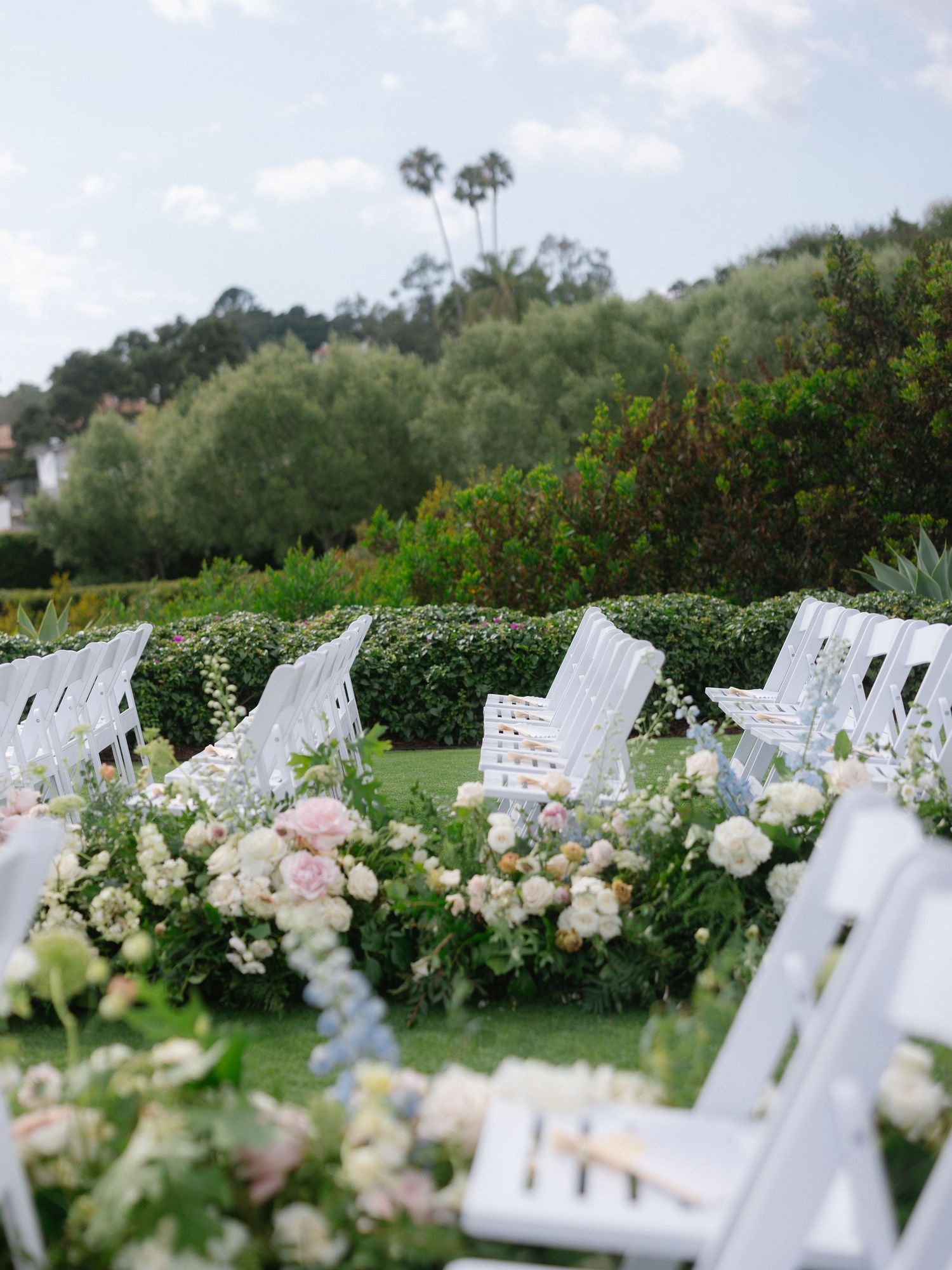 White garden chairs lined with lush floral arrangements of roses and greenery at outdoor ceremony.