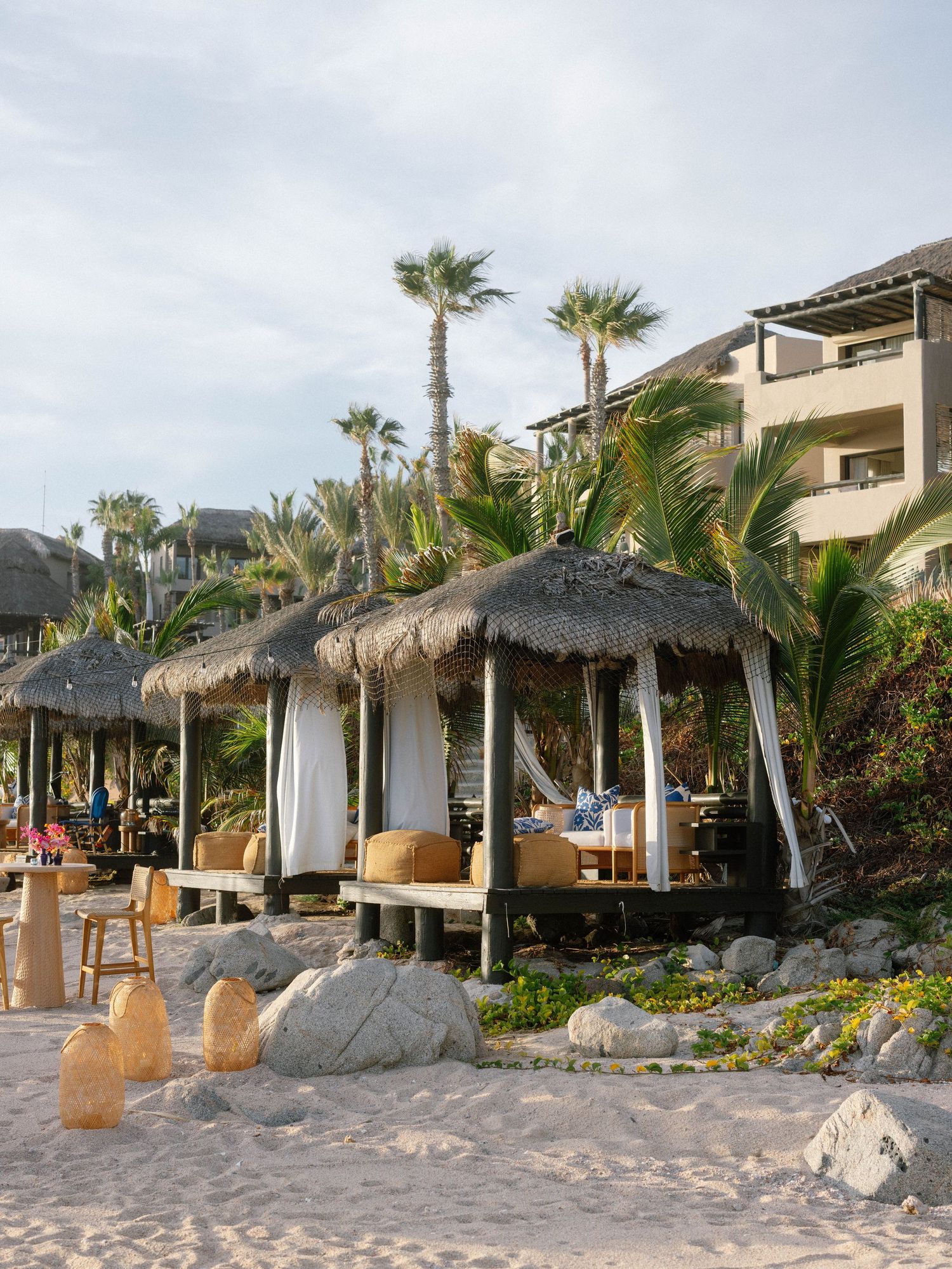 Tropical beach cabanas with thatched roofs, white curtains, and palm trees at a luxury beachfront resort.