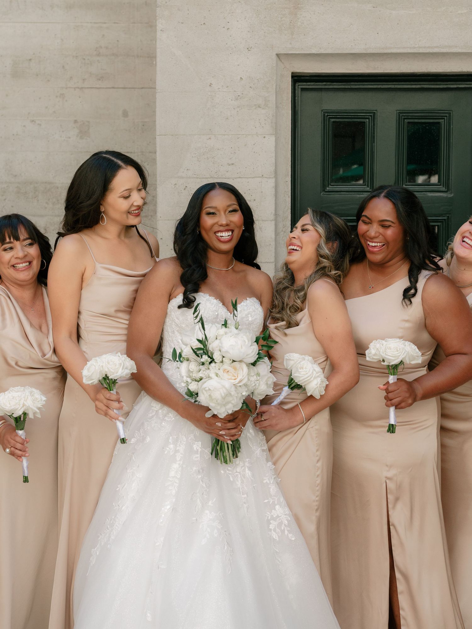 Smiling bride with bridesmaids in champagne gowns holding white bouquets outside stone venue.
