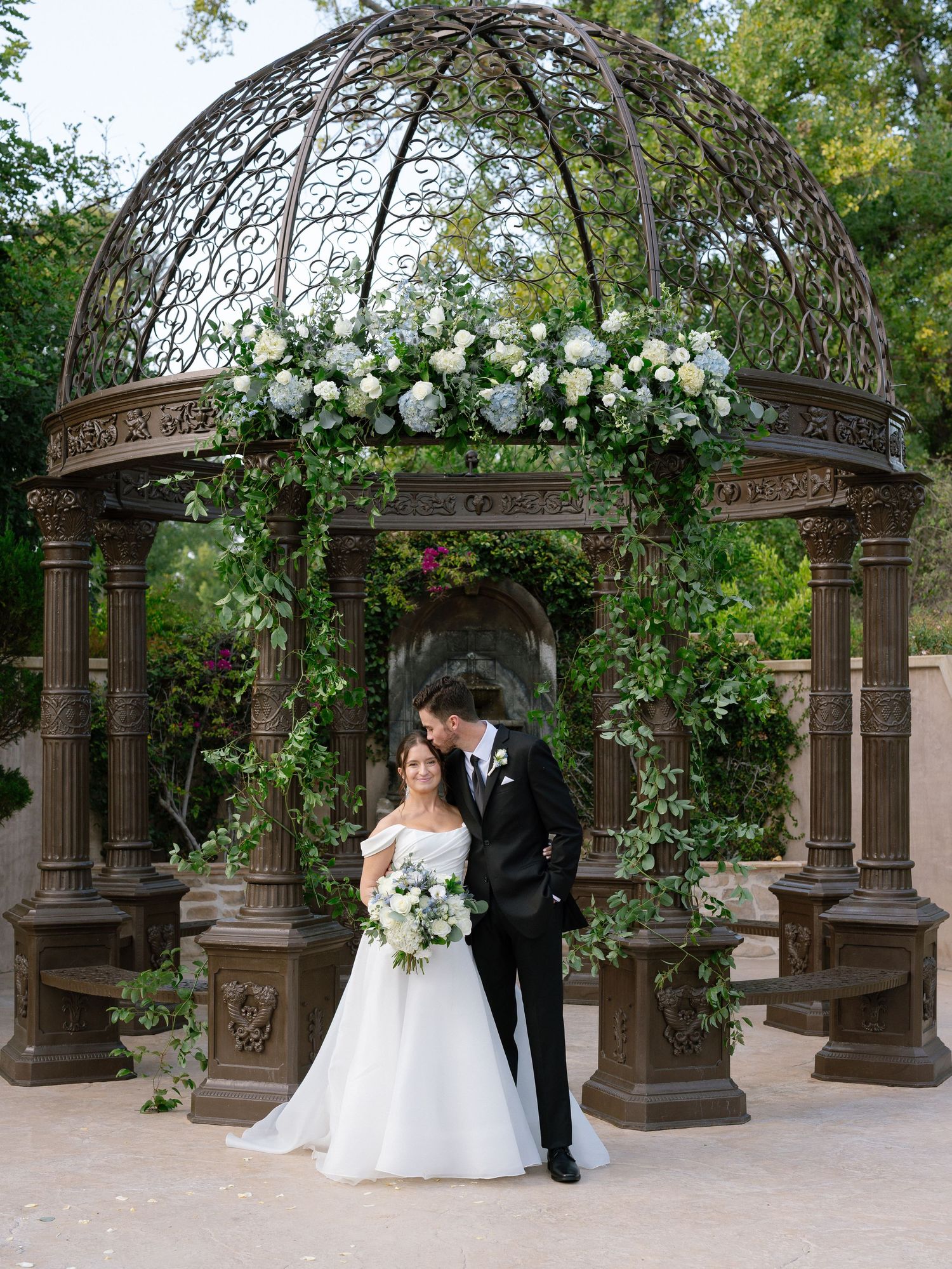 Bride and groom kissing under ornate iron gazebo decorated with white roses and lush green foliage.