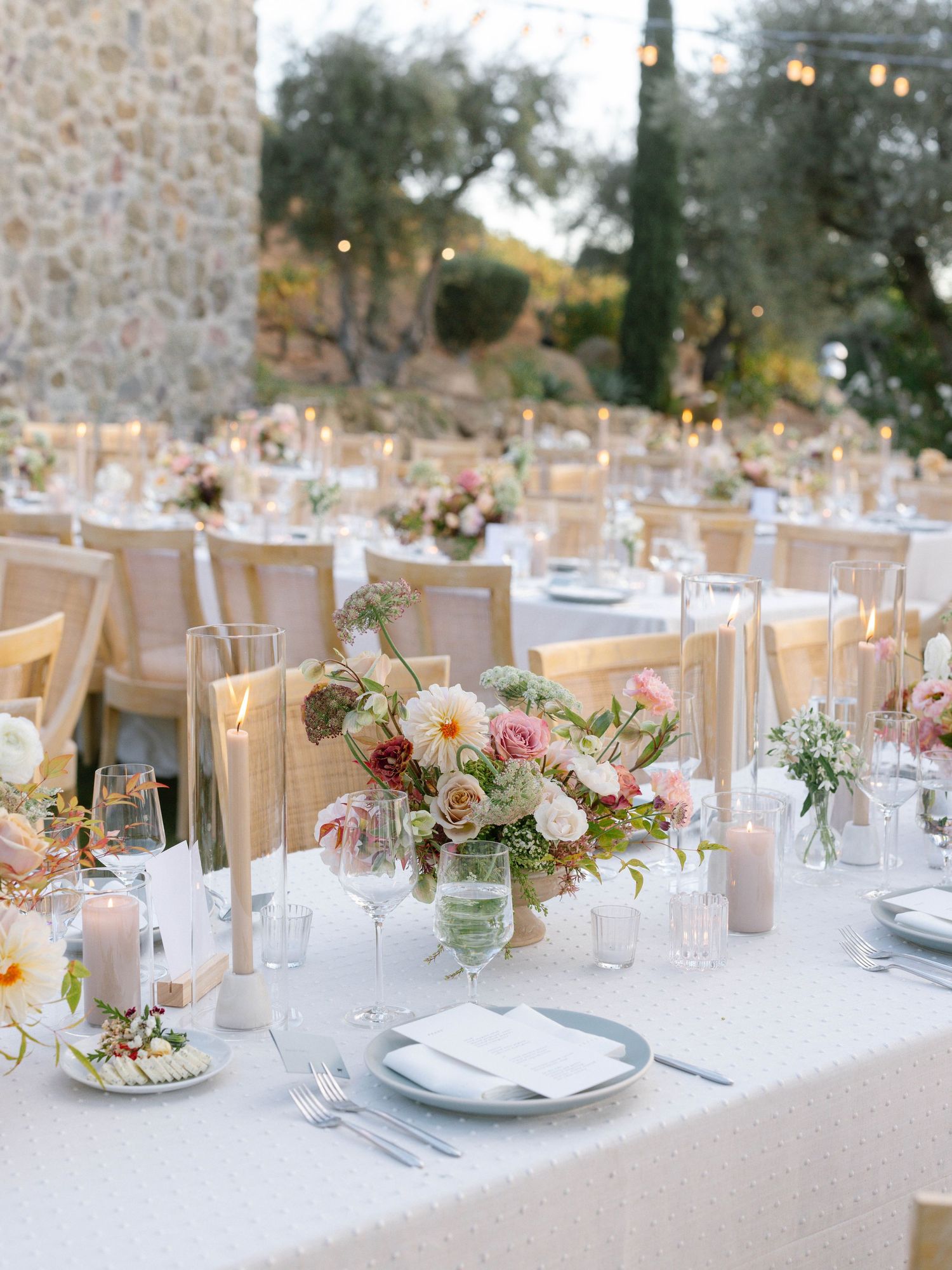 Elegant outdoor wedding reception with long tables, candles, blush floral centerpieces, and warm string lights.