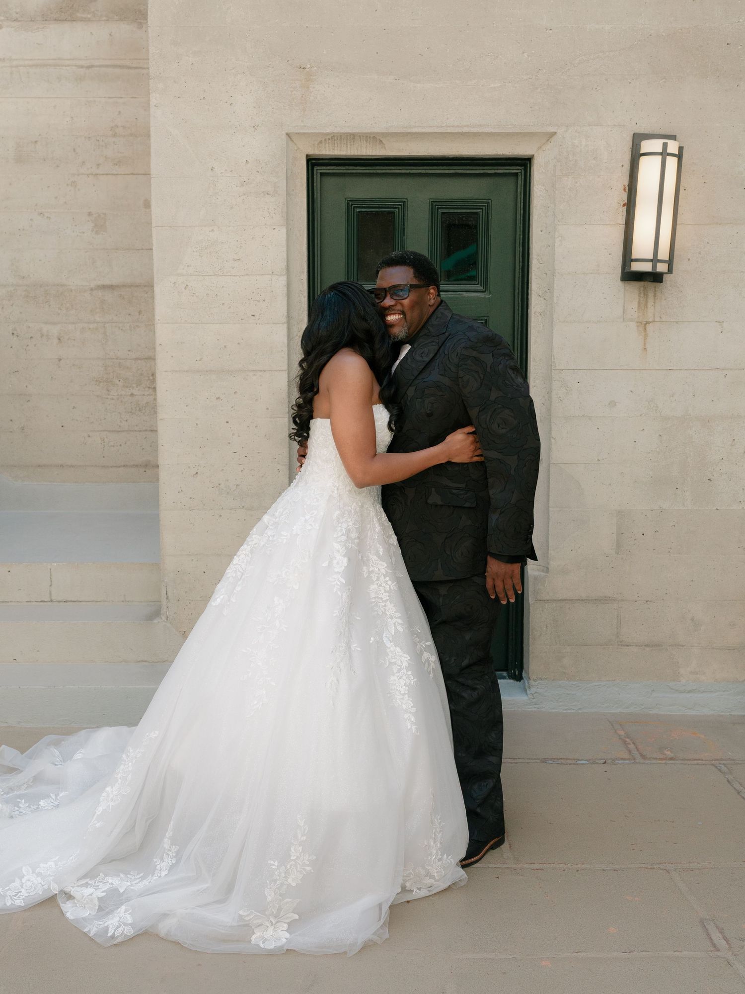 Bride in lace ball gown embraces guest in black suit outside stone wedding venue.