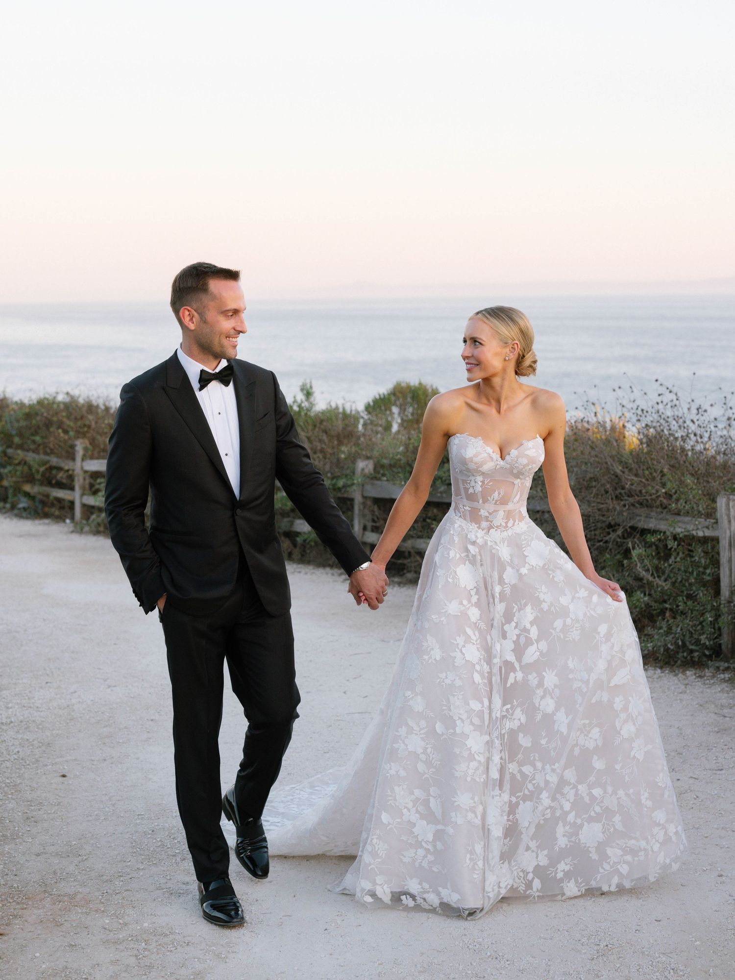 Bride and groom walk hand in hand along coastal path at dusk, bride in floral strapless ball gown.