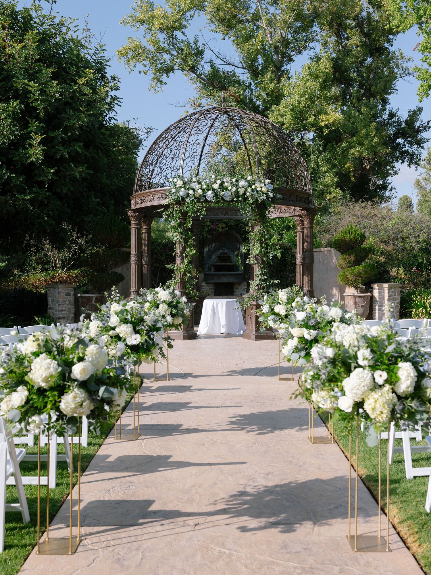Elegant outdoor ceremony aisle lined with white hydrangea arrangements leading to ornate iron gazebo.