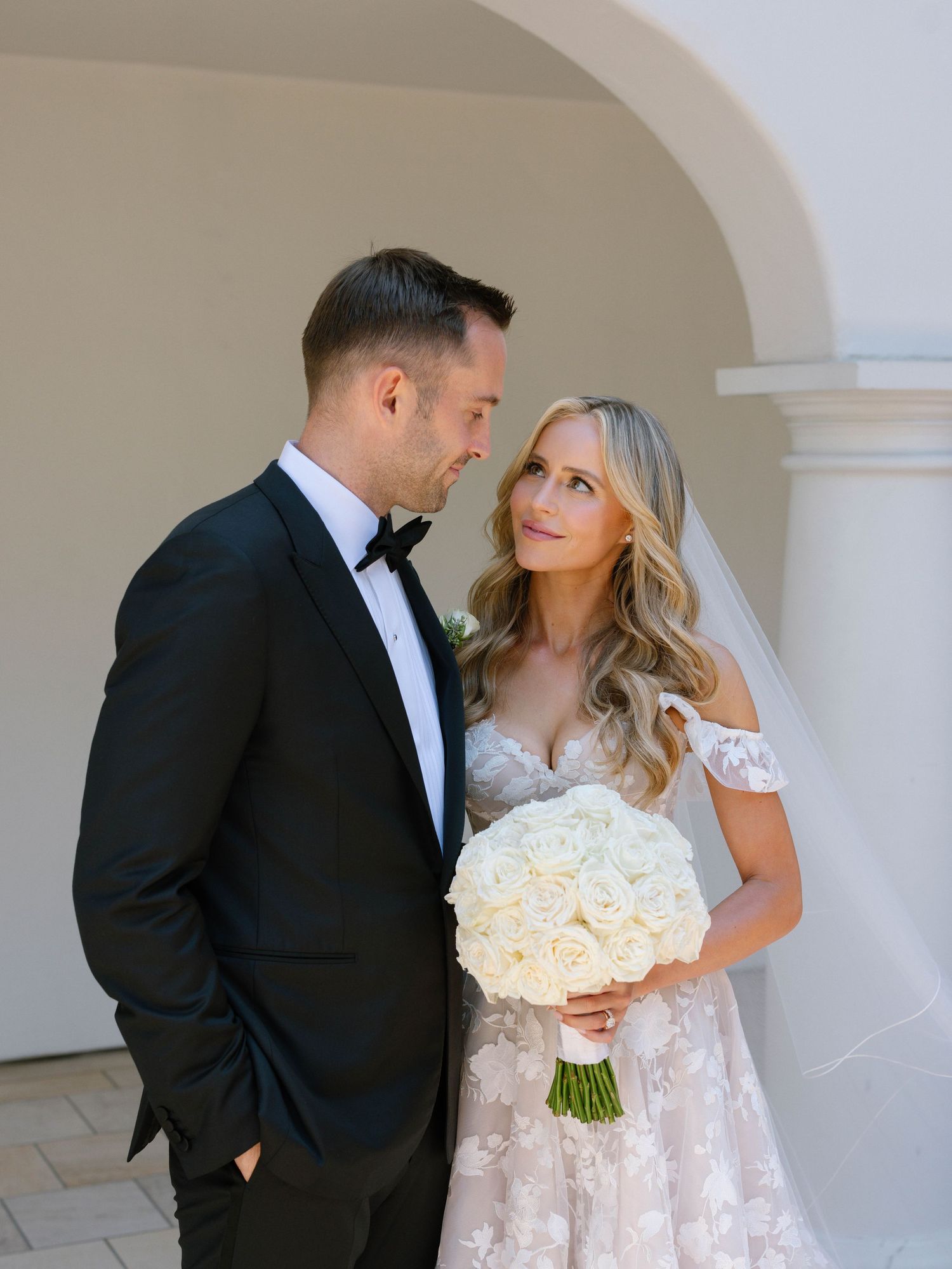 Bride and groom share a romantic moment, she holds white rose bouquet wearing lace off-shoulder gown, he wears black tuxedo.