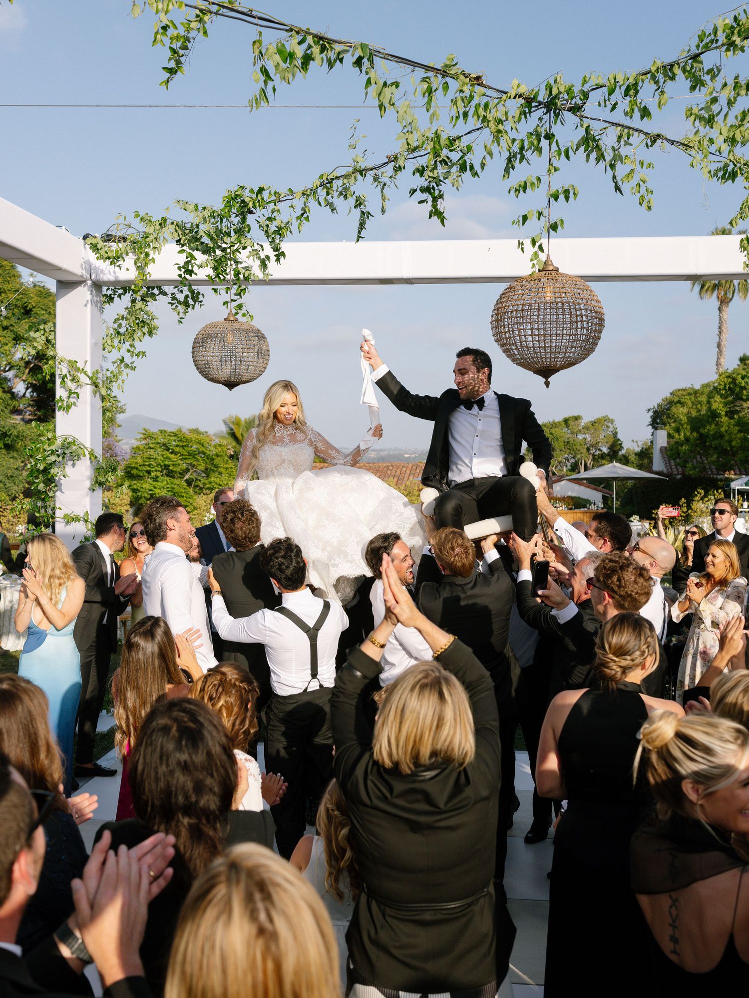 Bride and groom lifted by guests in chairs during joyful Jewish hora dance at outdoor wedding.