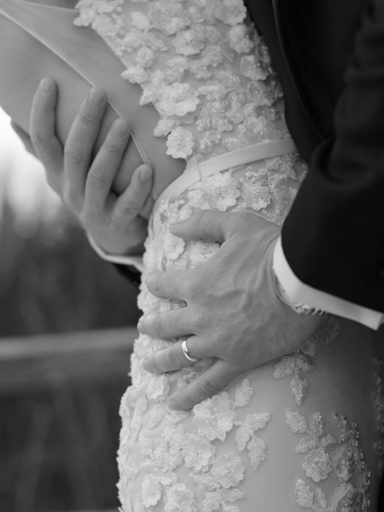 Black and white photo of bride and groom exchanging vows, groom's hand on bride's floral lace wedding dress.