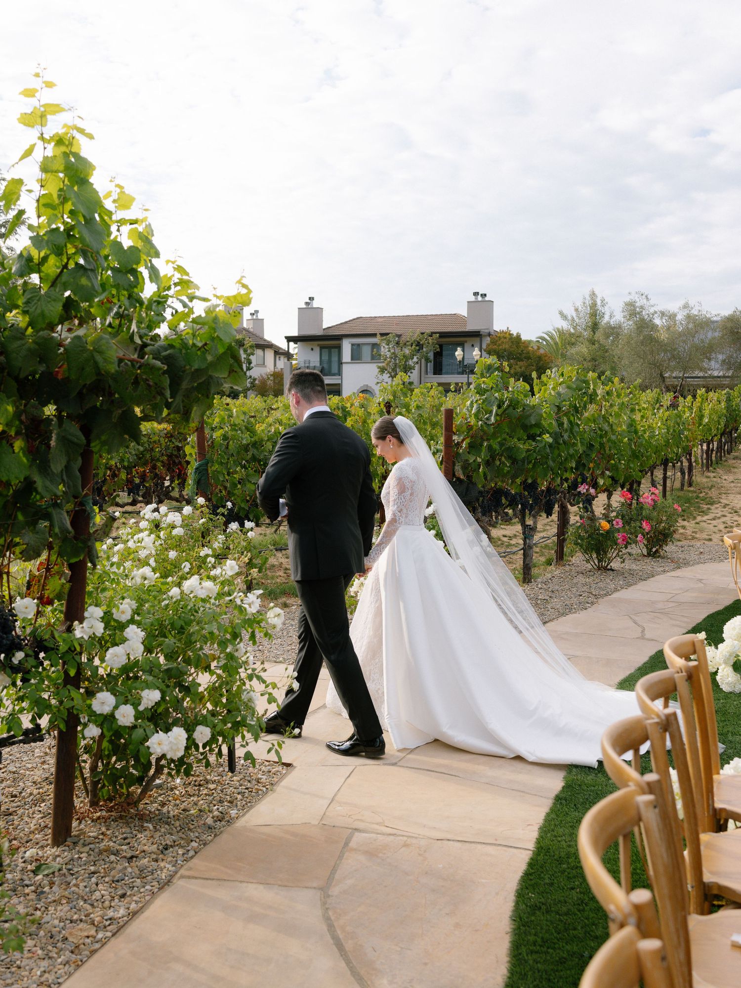 Bride and groom walk hand-in-hand through a scenic vineyard, her elegant gown trailing behind on a sunny wedding day in Napa.
