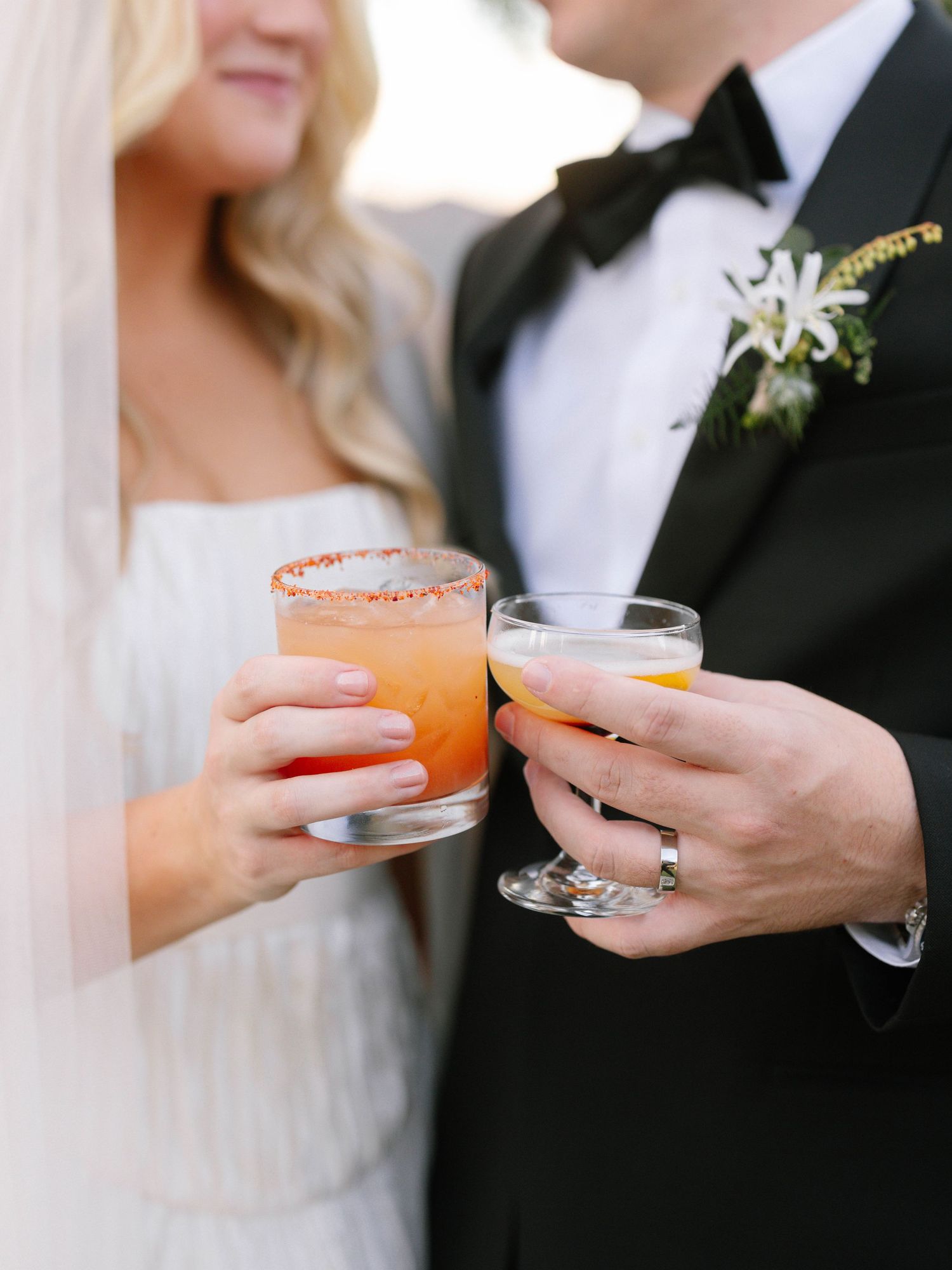 Bride and groom clink colorful cocktails, showcasing wedding rings and boutonniere at outdoor reception.