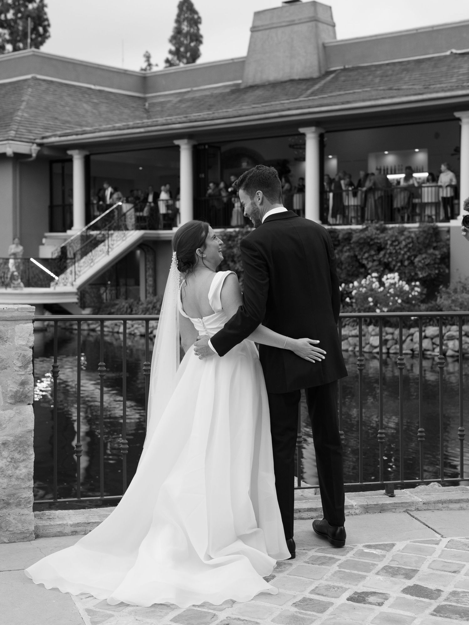 Black and white photo of bride and groom kissing near pond with wedding guests gathered on terrace behind.