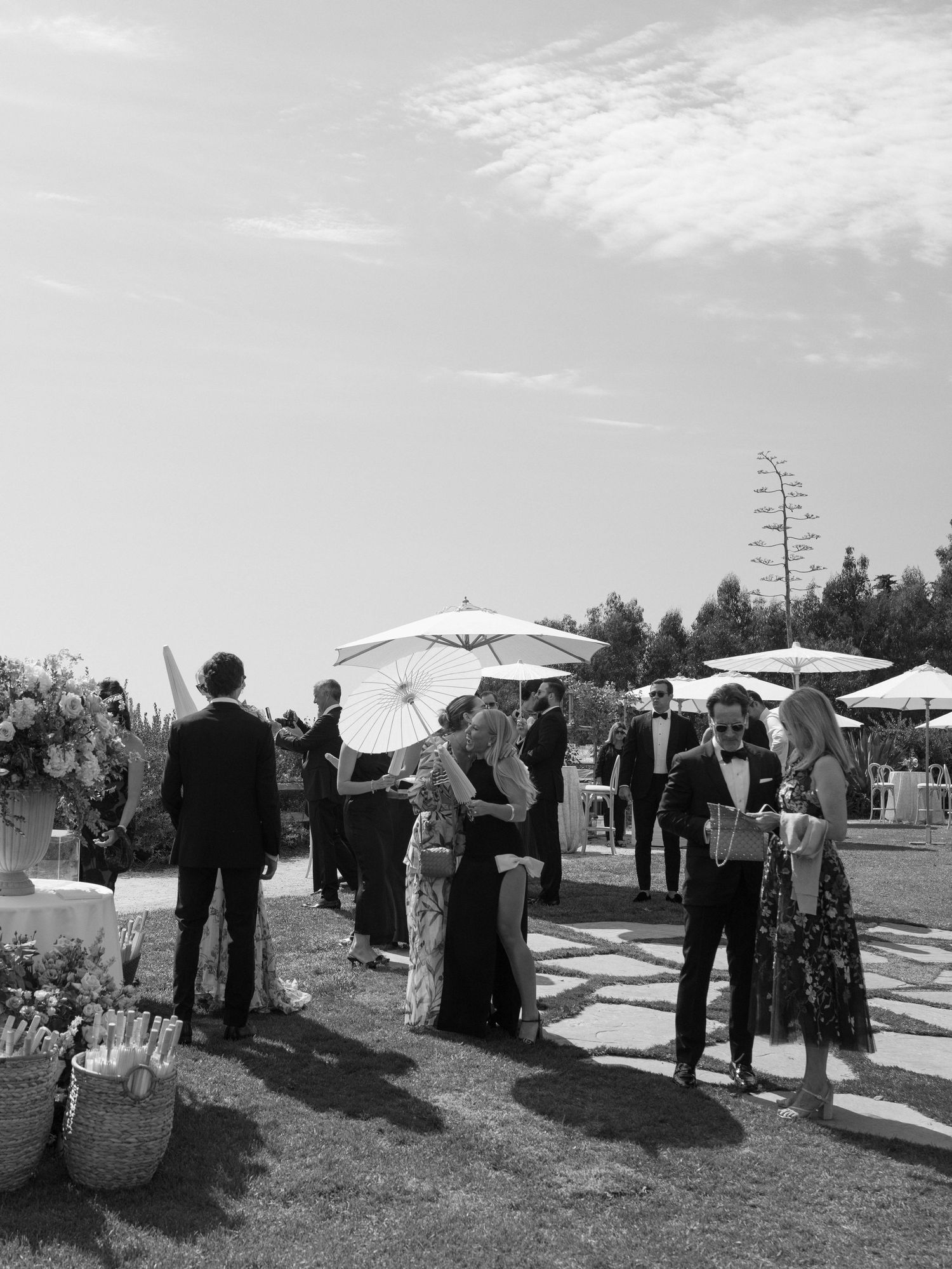 Black and white photo of wedding guests mingling outdoors under parasols at elegant garden cocktail hour.
