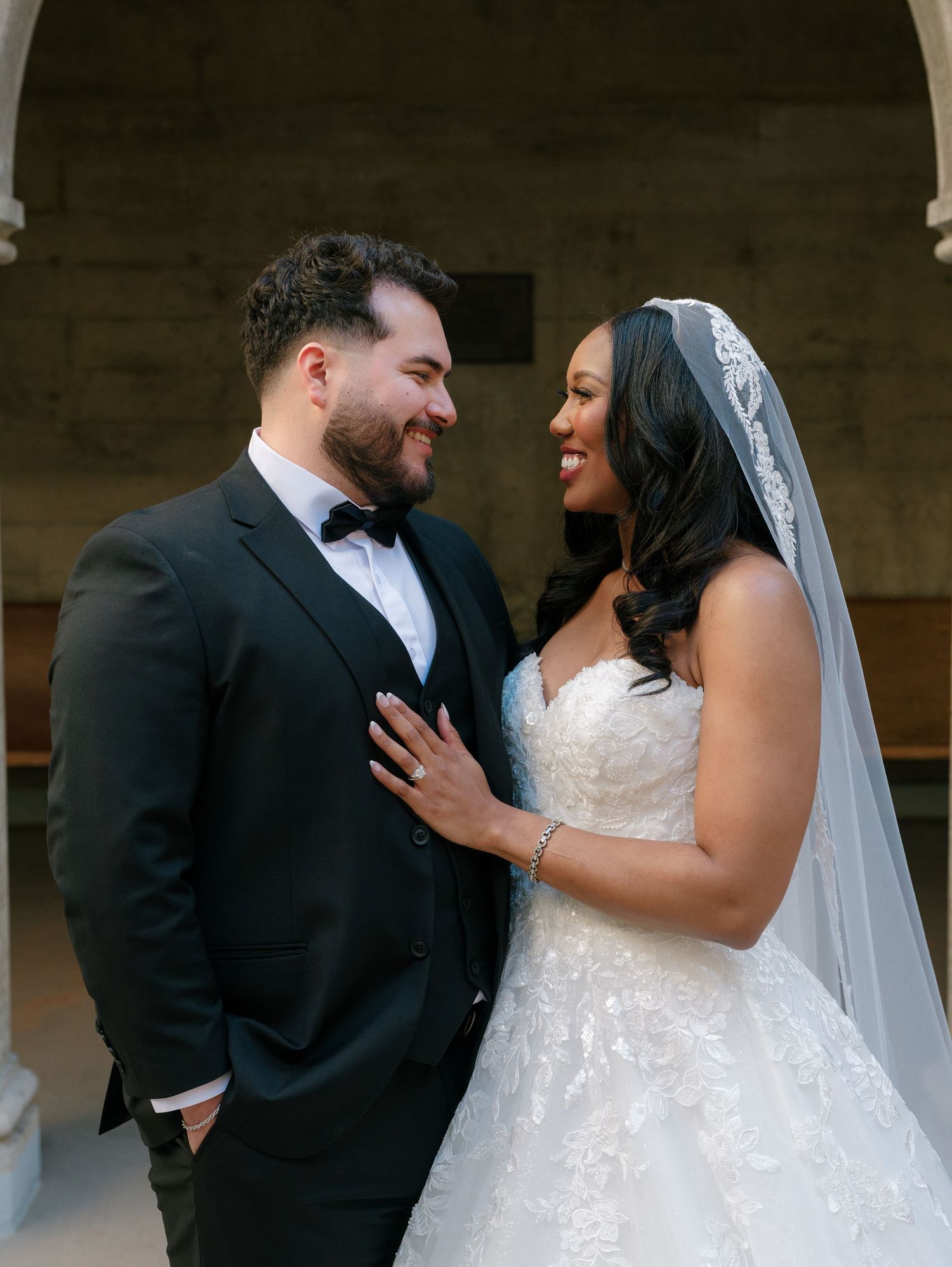 Interracial bride and groom share a joyful moment in front of stone archway at wedding.