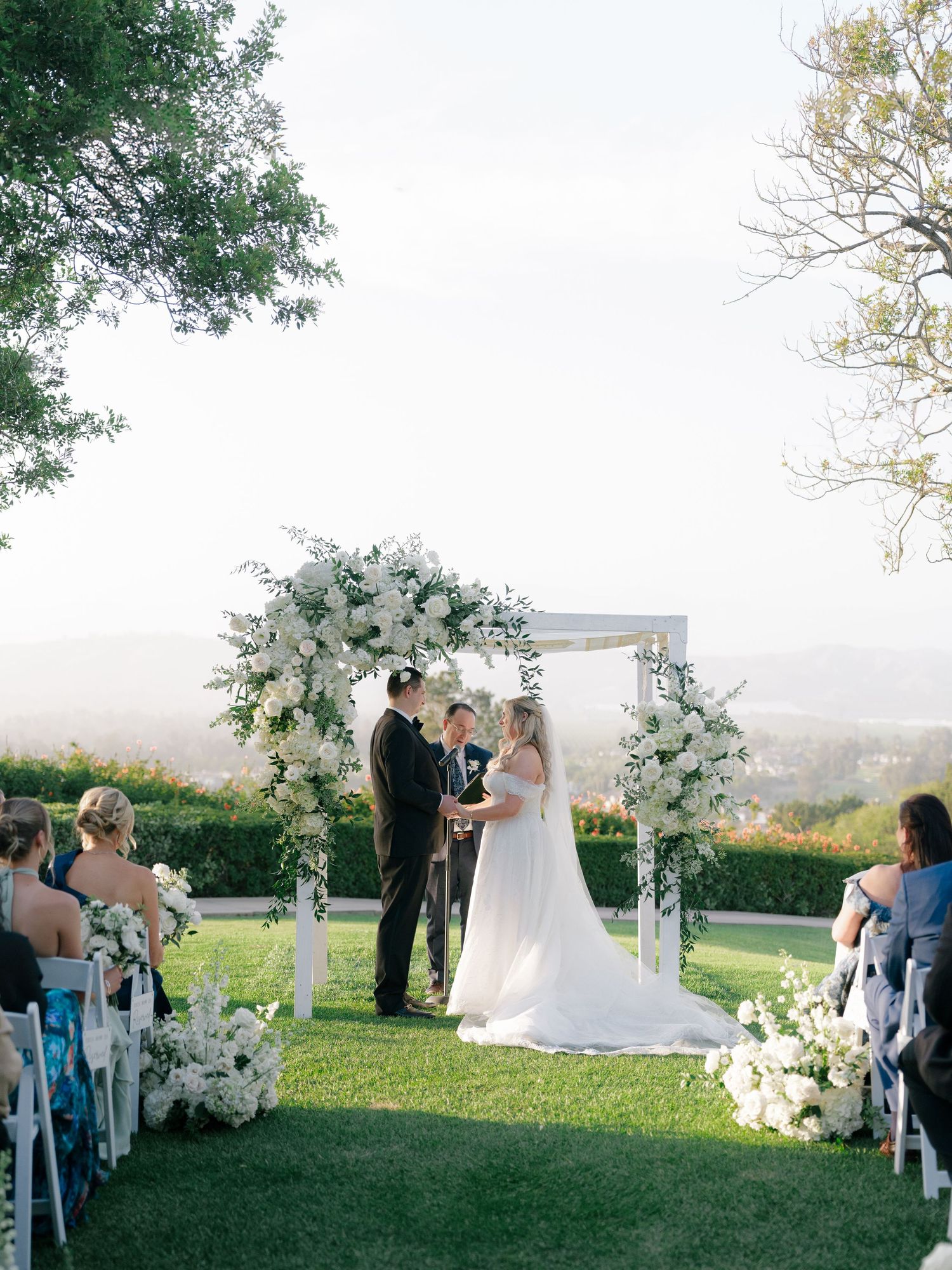 Outdoor wedding ceremony under white floral arch with white chairs on green lawn and scenic hillside view.