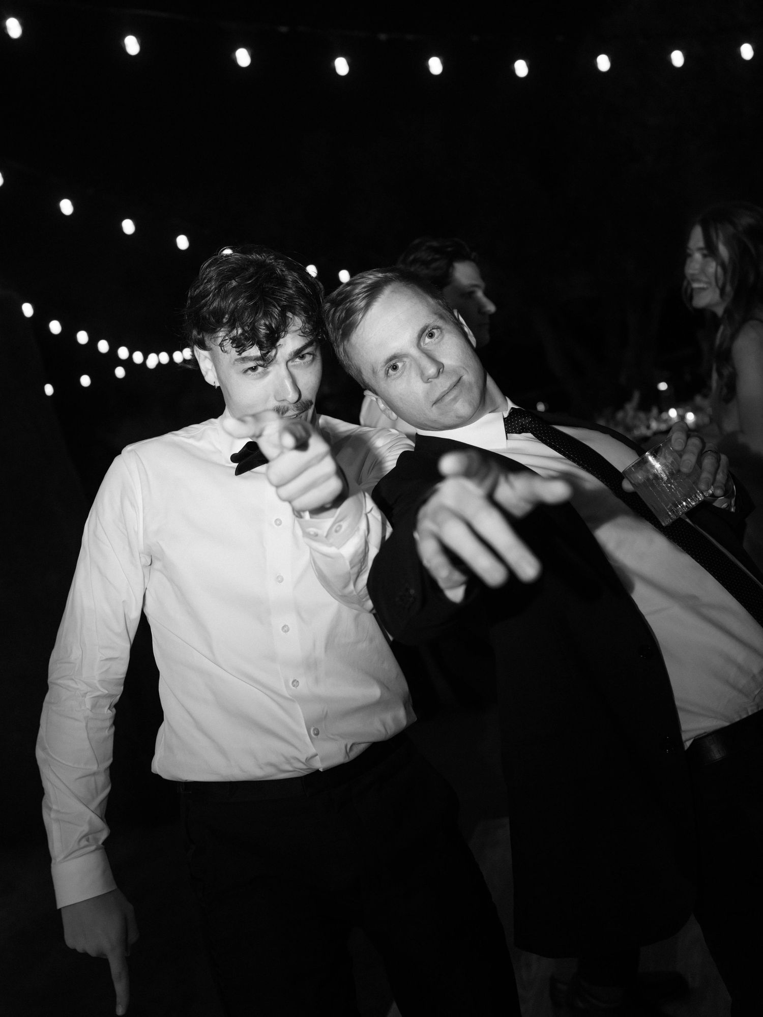 Two male wedding guests pointing at camera while dancing under string lights at evening reception.