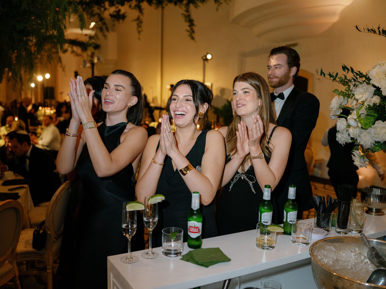 Three bridesmaids in black dresses clapping and cheering at a wedding reception bar.