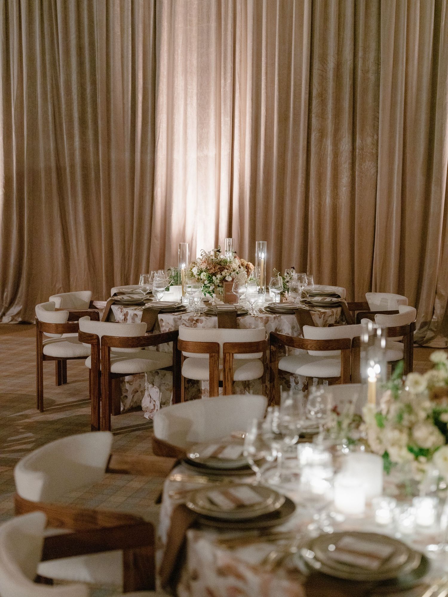 Elegant wedding reception round tables with floral centerpieces and white chairs against draped champagne curtains.