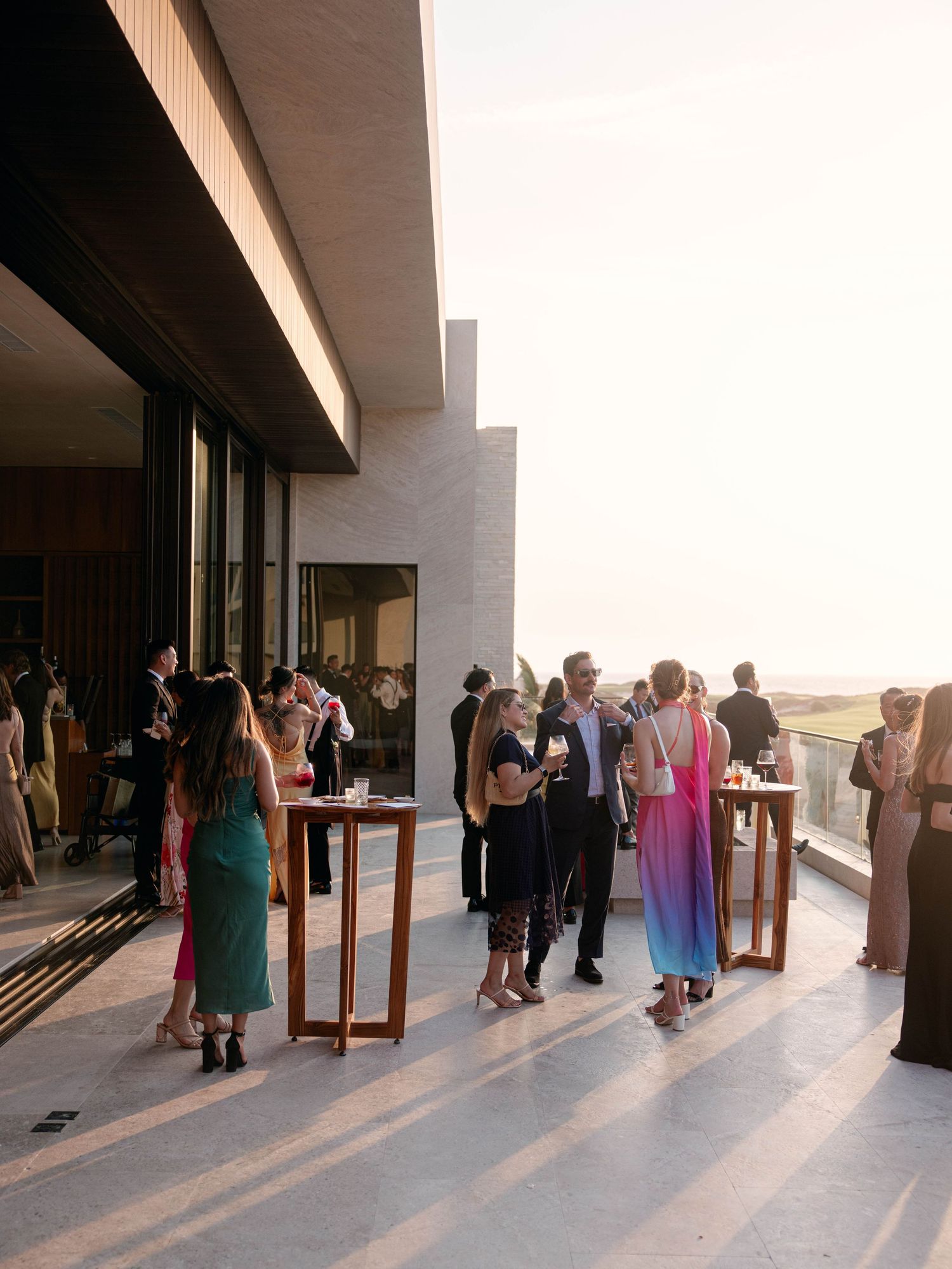 Wedding cocktail hour guests mingle on a modern rooftop terrace bathed in warm golden sunset light.