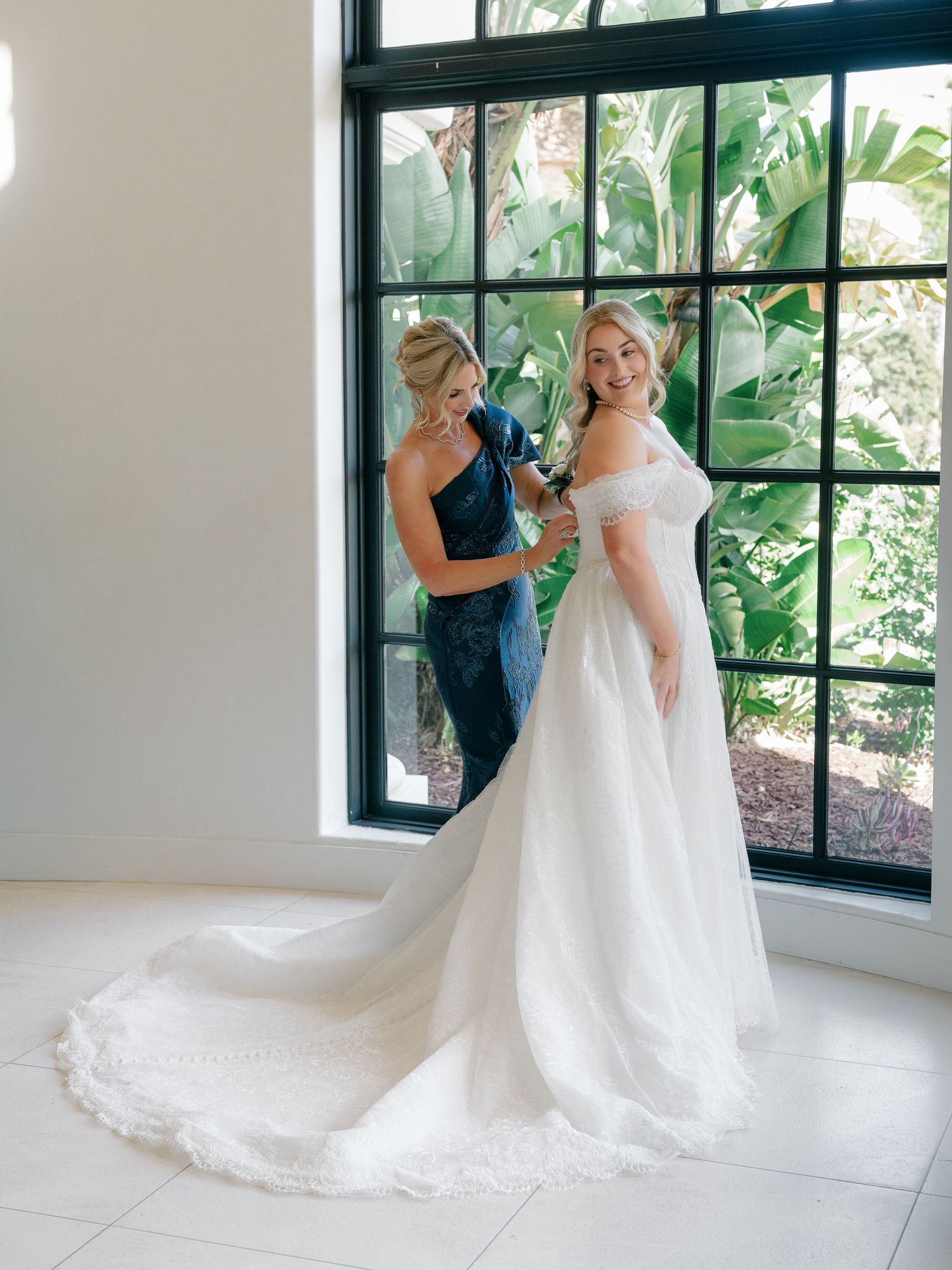 Bride in off-shoulder wedding gown with long train being helped by bridesmaid near large garden window.
